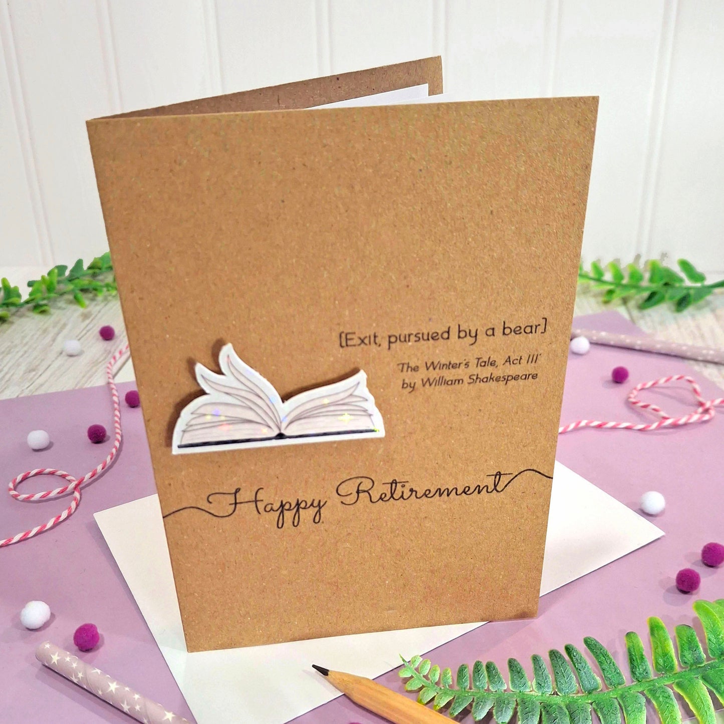 A brown kraft retirement card with illustrated book pages in a raised, holographic embellishment, the words Happy Retirement, and a quote from William Shakespeare's The Winter's Tale Act III. The card is displayed on a purple surface with decorative greenery, beads, and stationery.