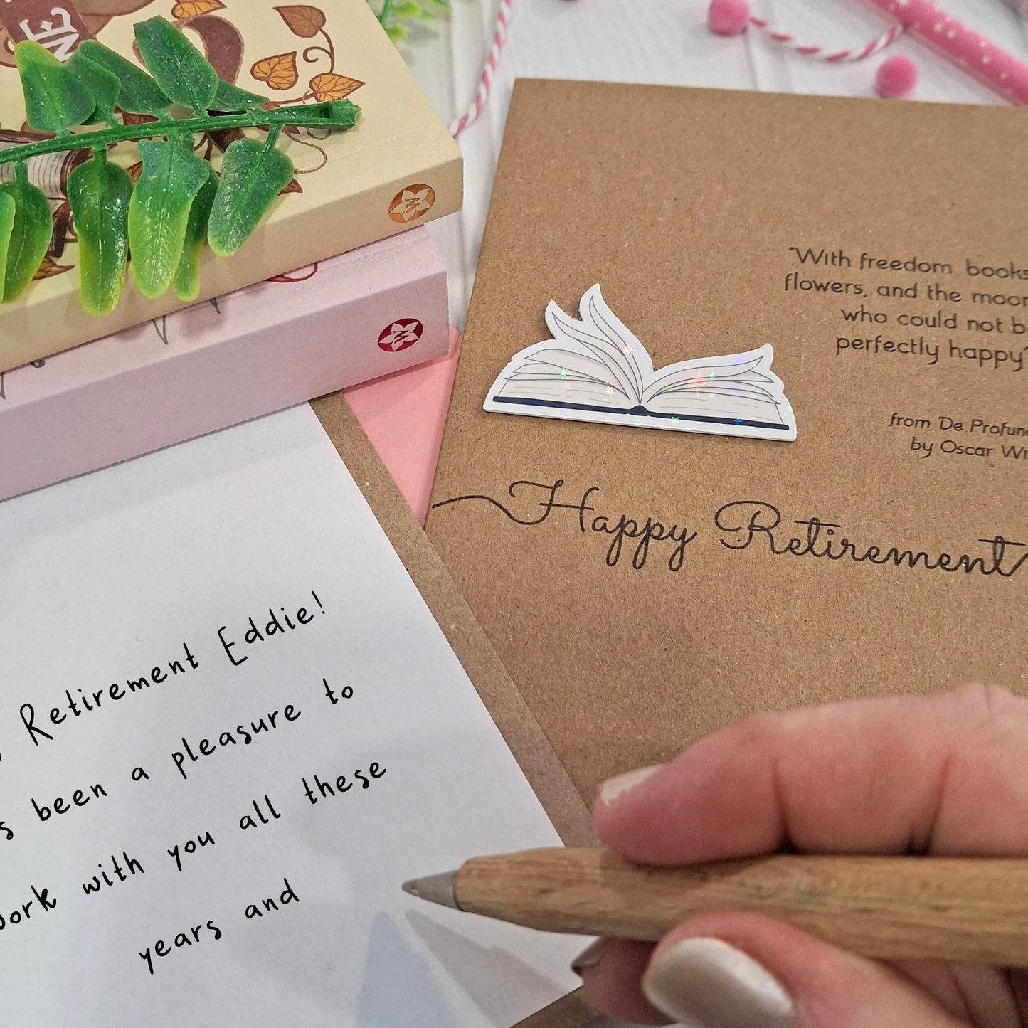 A close up of a hand holding a wooden pen, and writing a happy retirement message inside a card. The card features a raised holographic embellishment of an open book with gently fanned pages, the words Happy Retirement, and a quote by Oscar Wilde.