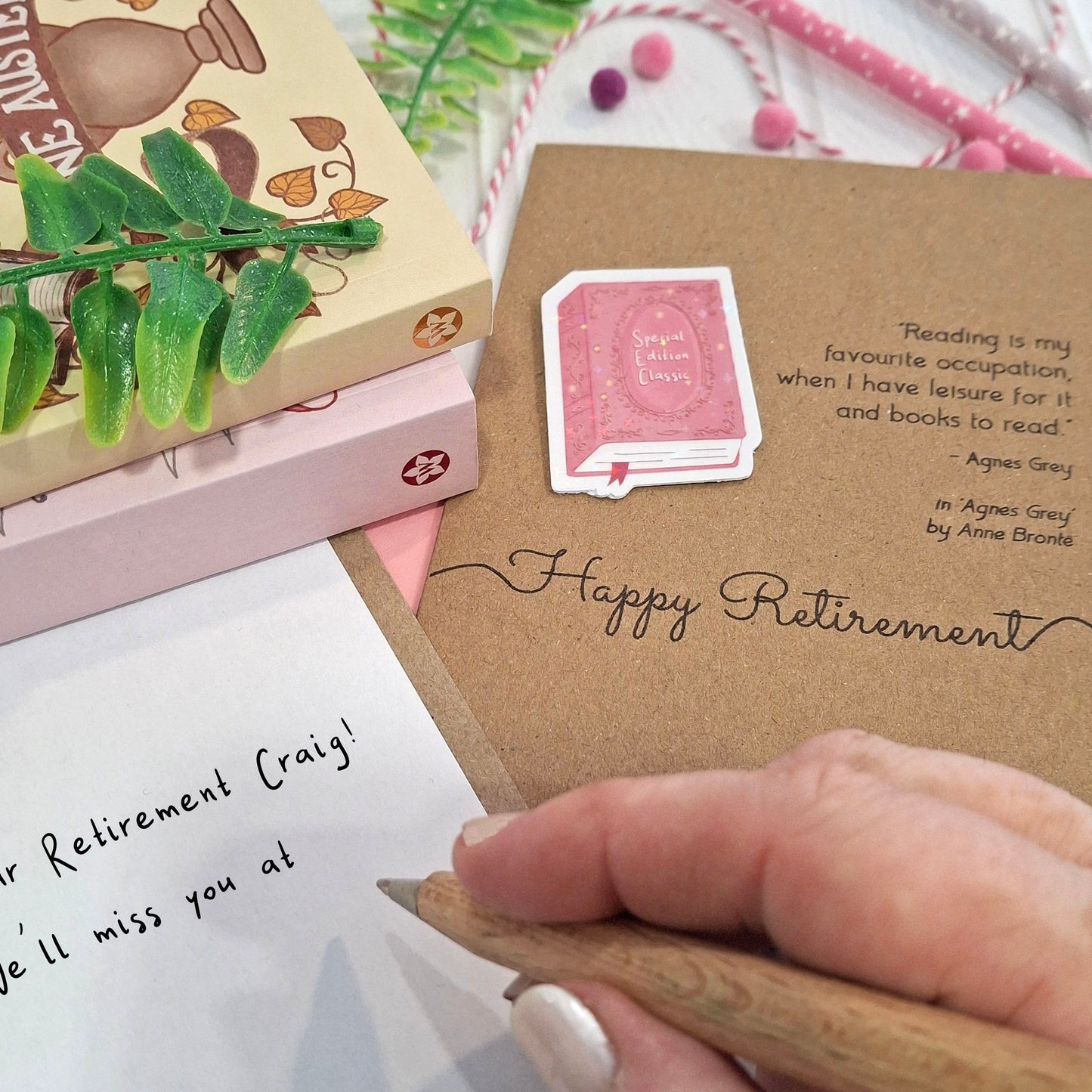 A close-up of a hand holding a wooden pen and writing a retirement message inside a card. The card features a raised holographic embellishment of a pink special edition classic book, the words Happy Retirement, and a quote from Agnes Grey by Anne Bronte.