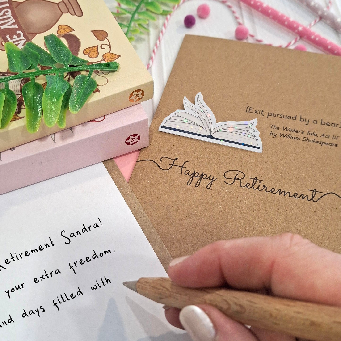 A close up of a hand holding a wooden pen, and writing a happy retirement message inside a card. The card features a raised holographic embellishment of an open book with gently fanned pages, the words Happy Retirement, and a quote from The Winter's Tale by William Shakespeare.
