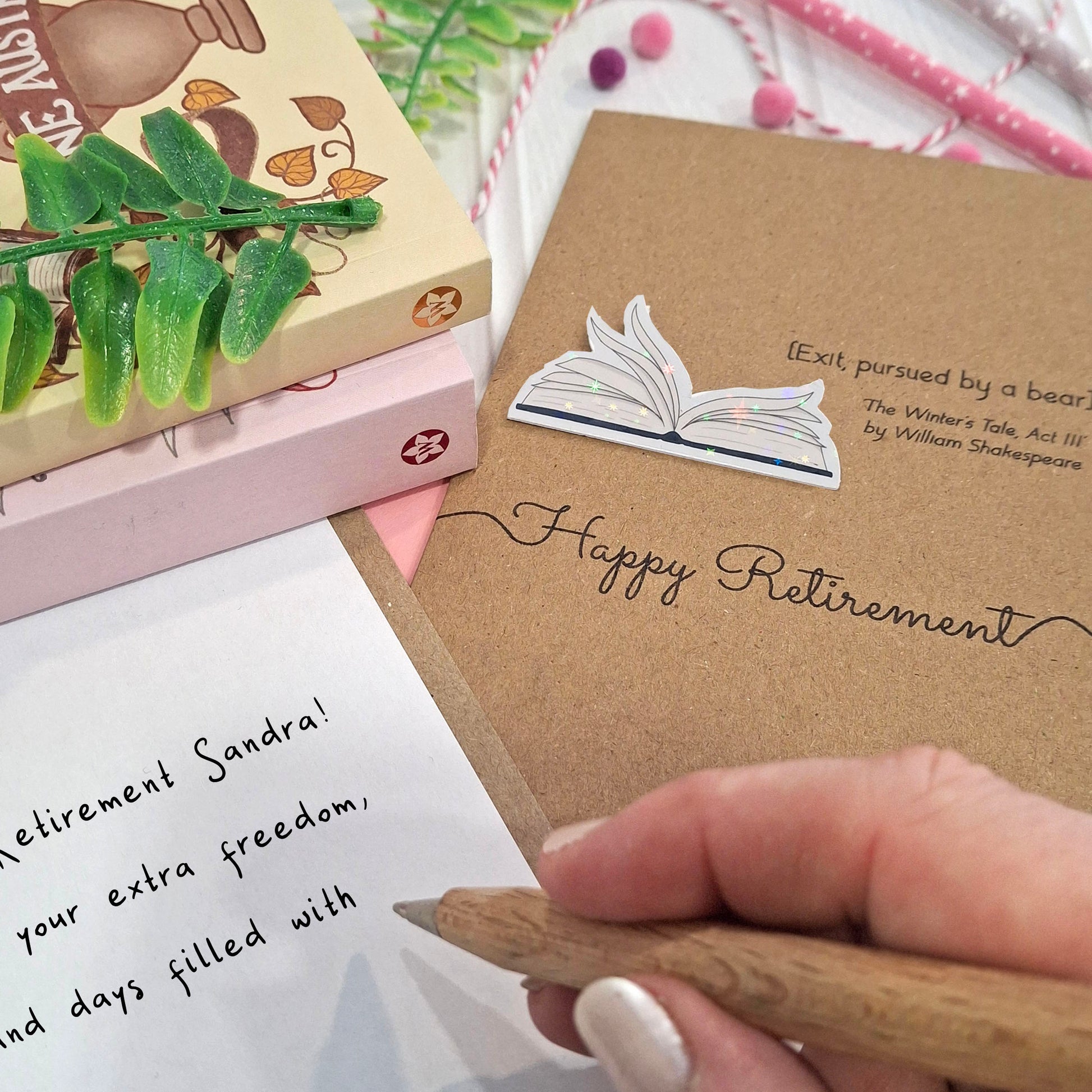 A close up of a hand holding a wooden pen, and writing a happy retirement message inside a card. The card features a raised holographic embellishment of an open book with gently fanned pages, the words Happy Retirement, and a quote from The Winter's Tale by William Shakespeare.