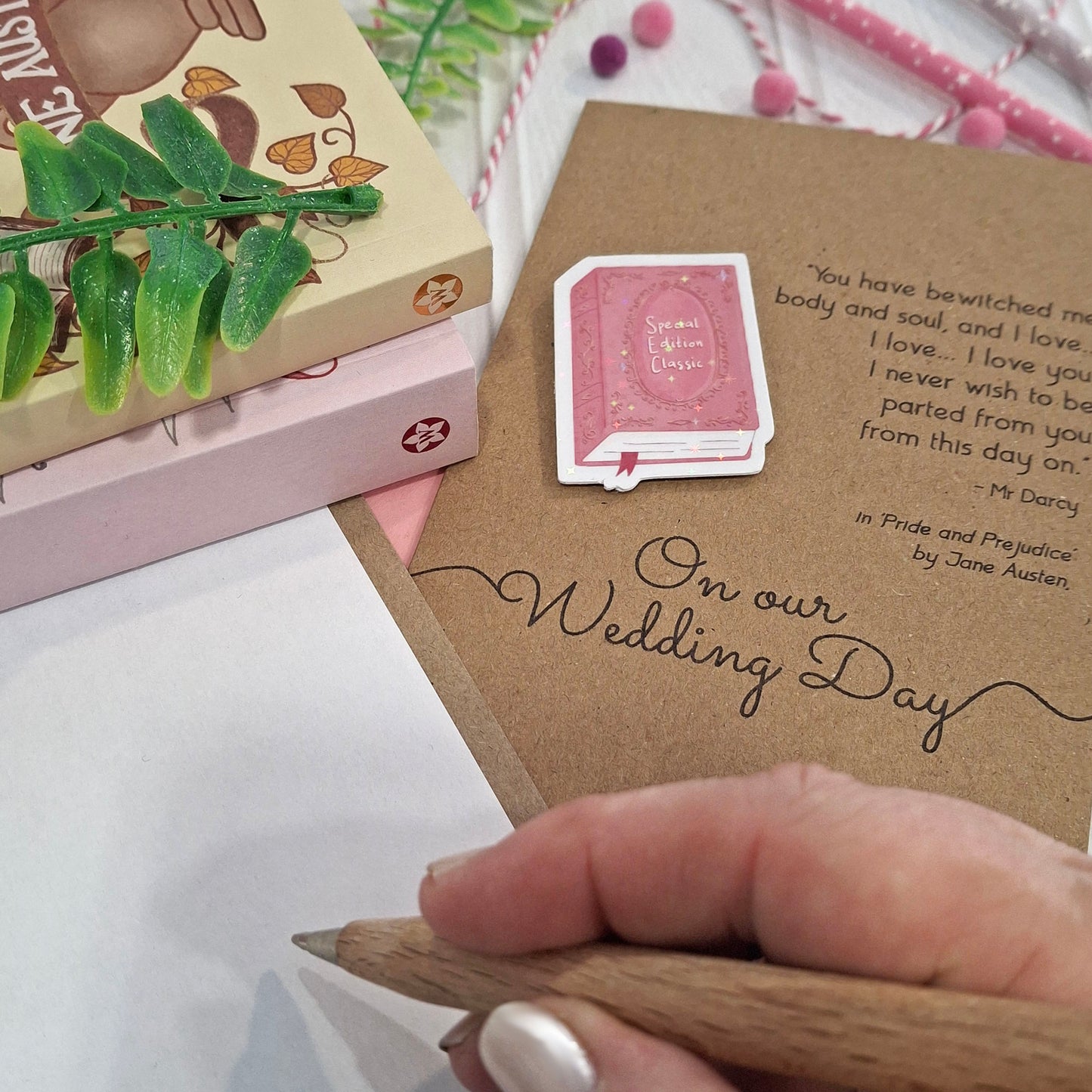 A close-up of a hand holding a wooden pencil, writing inside a card styled with decorative props. The card features a raised holographic embellishment of a pink special edition classic book, the words On Our Wedding Day, and a quote from Pride and Prejudice by Jane Austen.
