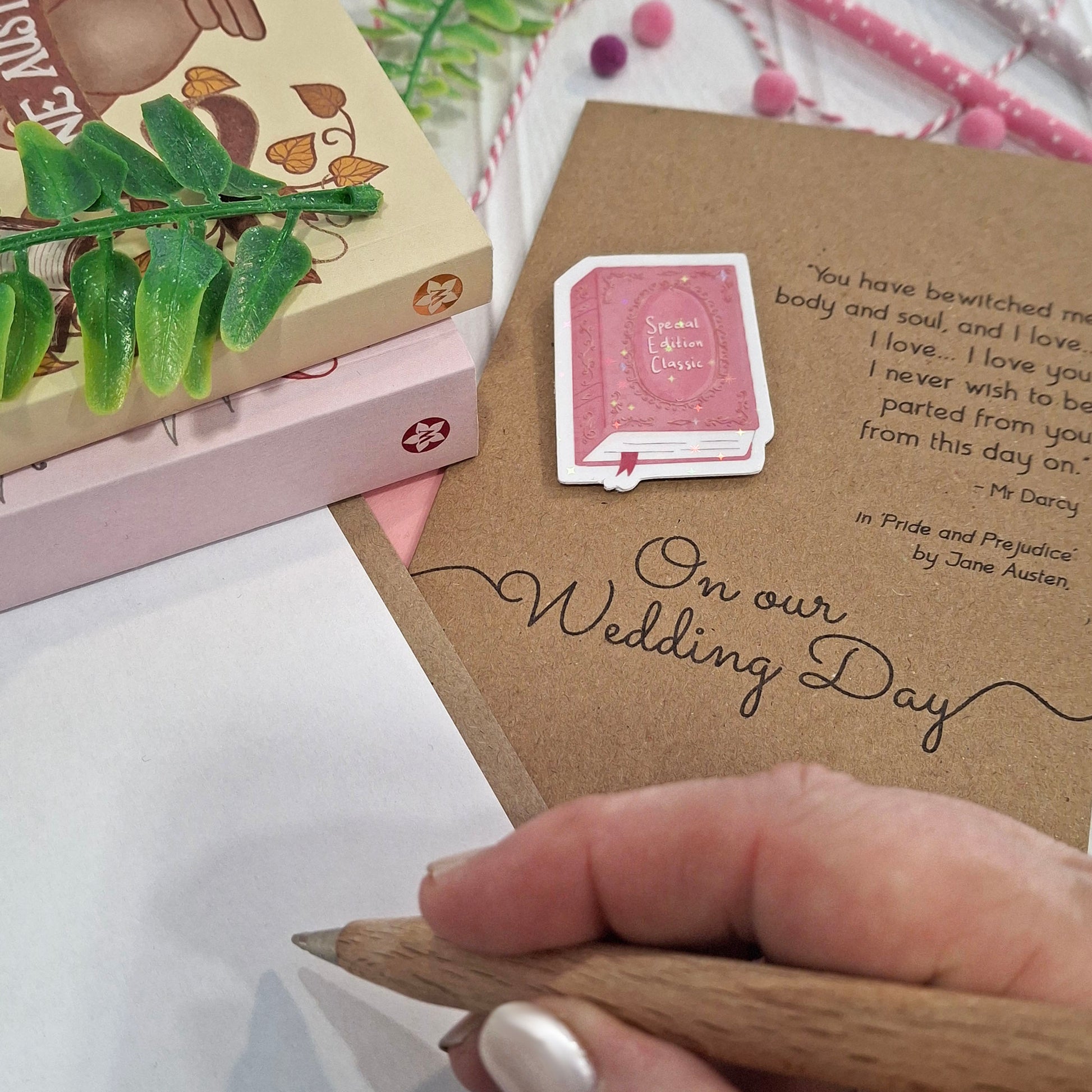 A close-up of a hand holding a wooden pencil, writing inside a card styled with decorative props. The card features a raised holographic embellishment of a pink special edition classic book, the words On Our Wedding Day, and a quote from Pride and Prejudice by Jane Austen.