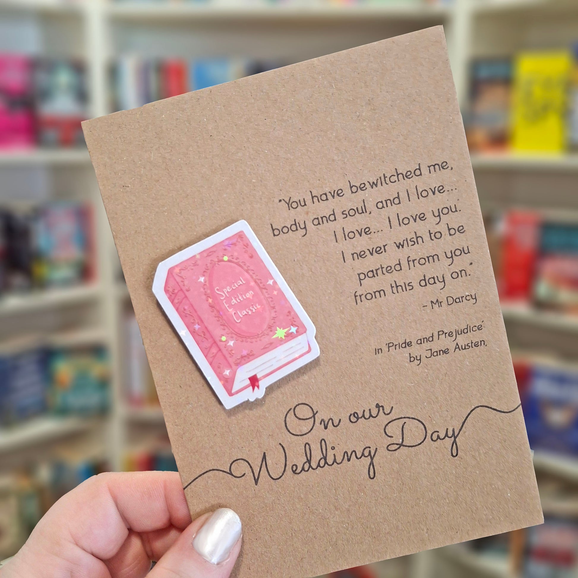 A hand holds a brown kraft Wedding Day card featuring a raised holographic embellishment of a pink special edition classic book, and a printed quote by Jane Austen. The background shows a blurred bookshop interior.