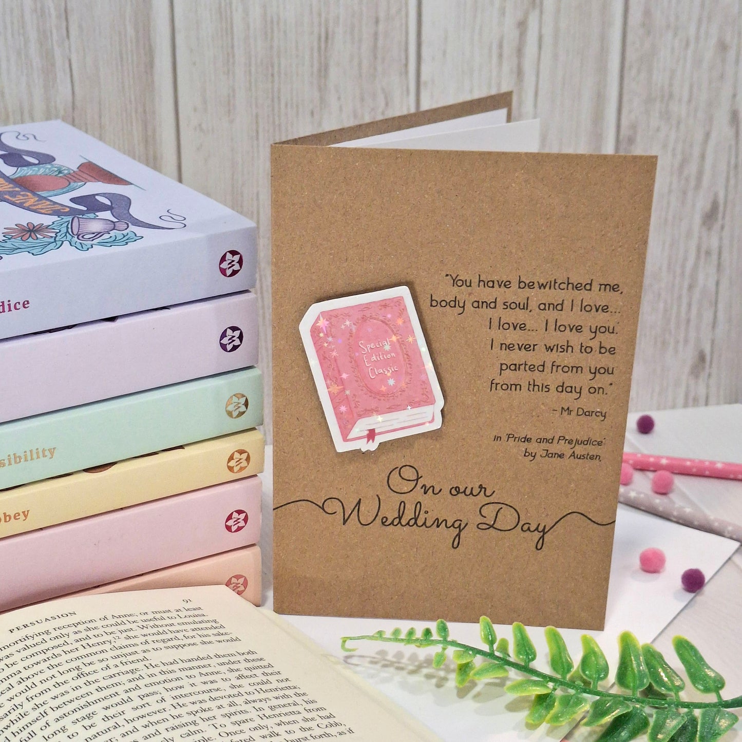 A kraft wedding card styled on a tabletop with books, an open book, and greenery. The card features a raised holographic embellishment of a pink special edition classic book, the words On Our Wedding Day, and a quote from Pride and Prejudice by Jane Austen.