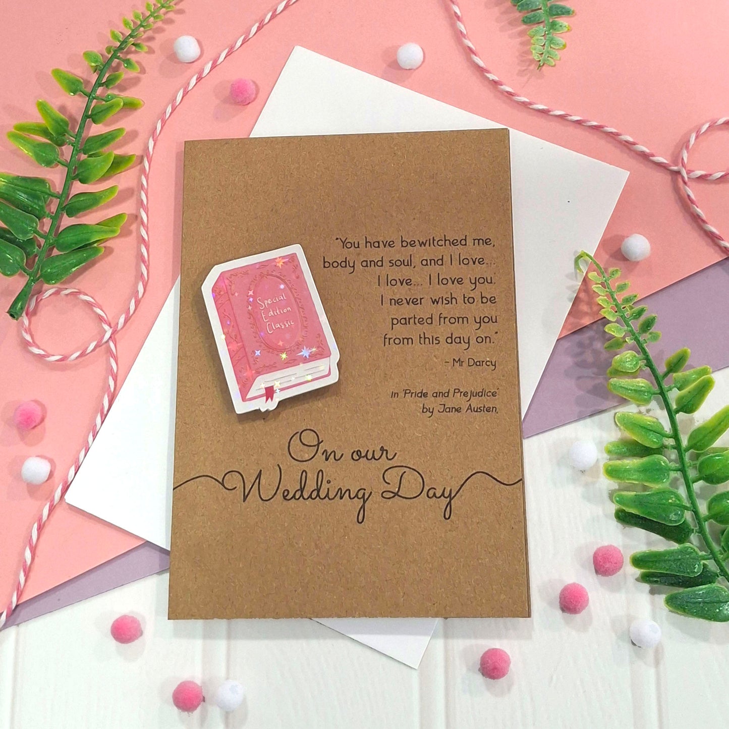 A kraft wedding card with a raised, holographic, illustrated story book embellishment in pink, a quote from Pride and Prejudice by Jane Austen, and the words On Your Wedding Day. The card is styled on a pink and white surface with white envelopes, string, faux greenery, and pink pom-poms.