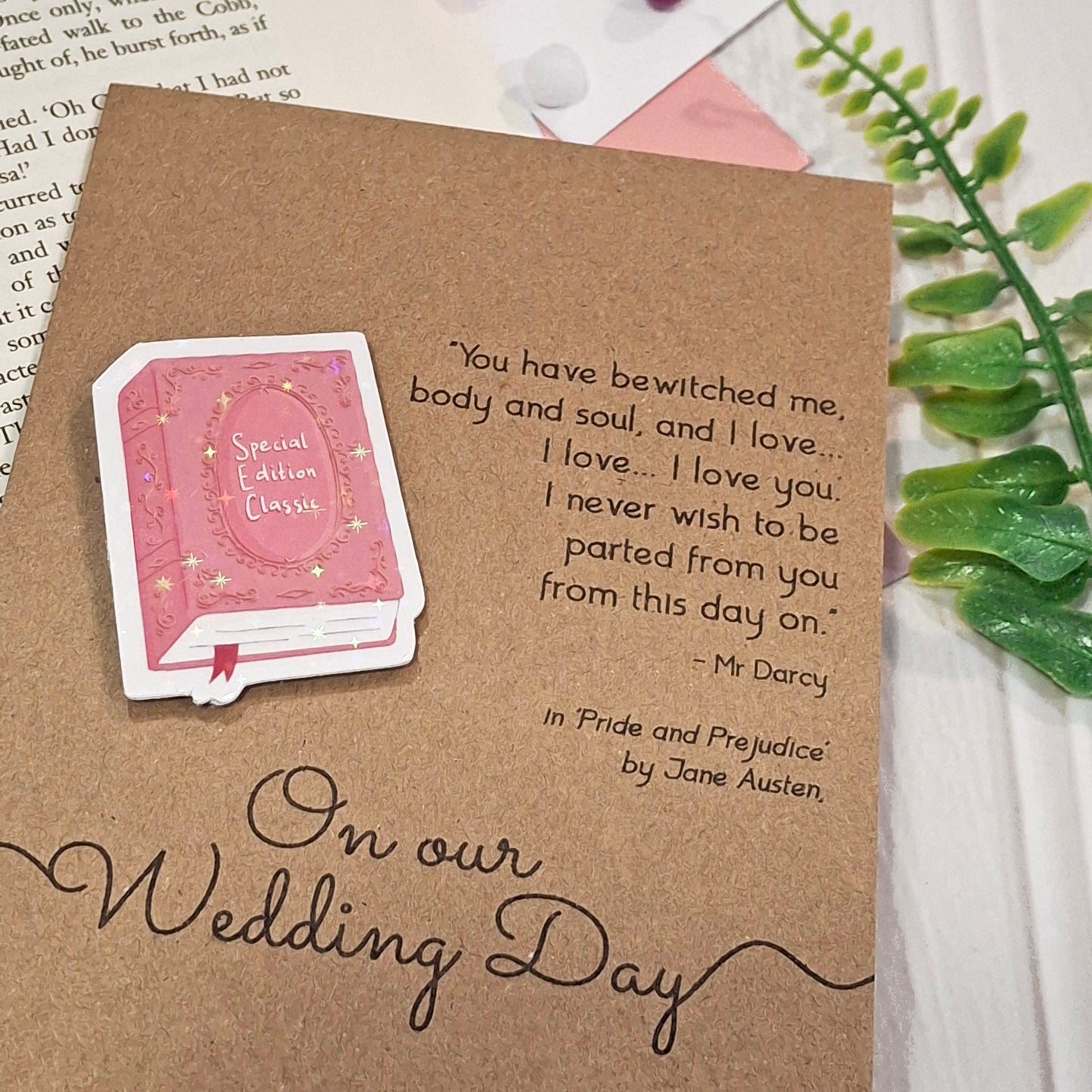 A kraft card styled on a white wood surface with a green leaf and book pages in the background. The card features a raised holographic embellishment of a pink special edition classic book, the words On Our Wedding Day, and a quote from Pride and Prejudice by Jane Austen.