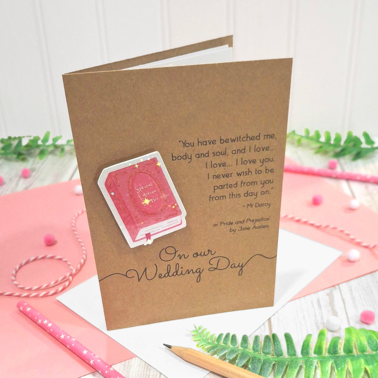 A kraft wedding card with a raised, holographic, illustrated story book embellishment in pink, a quote from Pride and Prejudice by Jane Austen, and the words On Our Wedding Day, standing on a pink and white tabletop with a pencil and greenery.