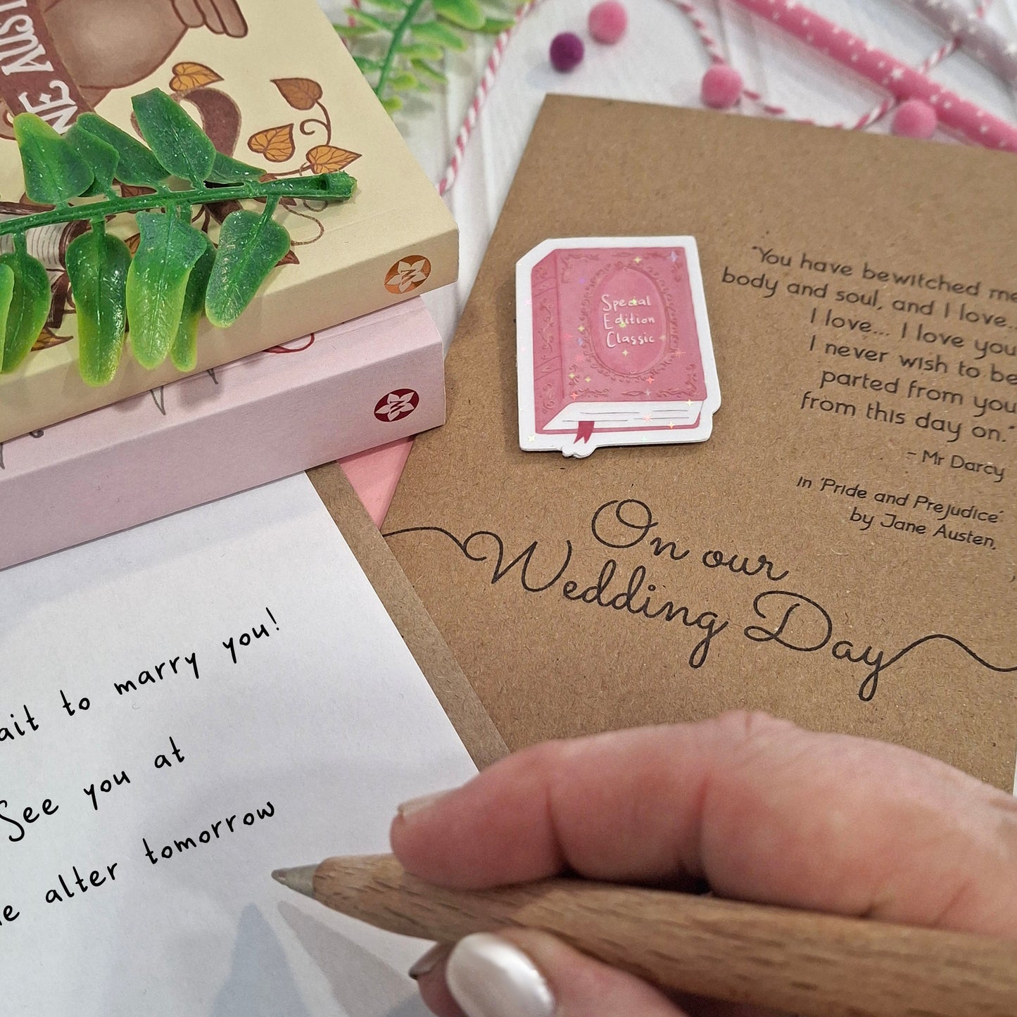 A close-up of a hand holding a wooden pencil, writing a love message inside a card, styled with decorative props. The card features a raised holographic embellishment of a pink special edition classic book, the words On Our Wedding Day, and a quote from Pride and Prejudice by Jane Austen.