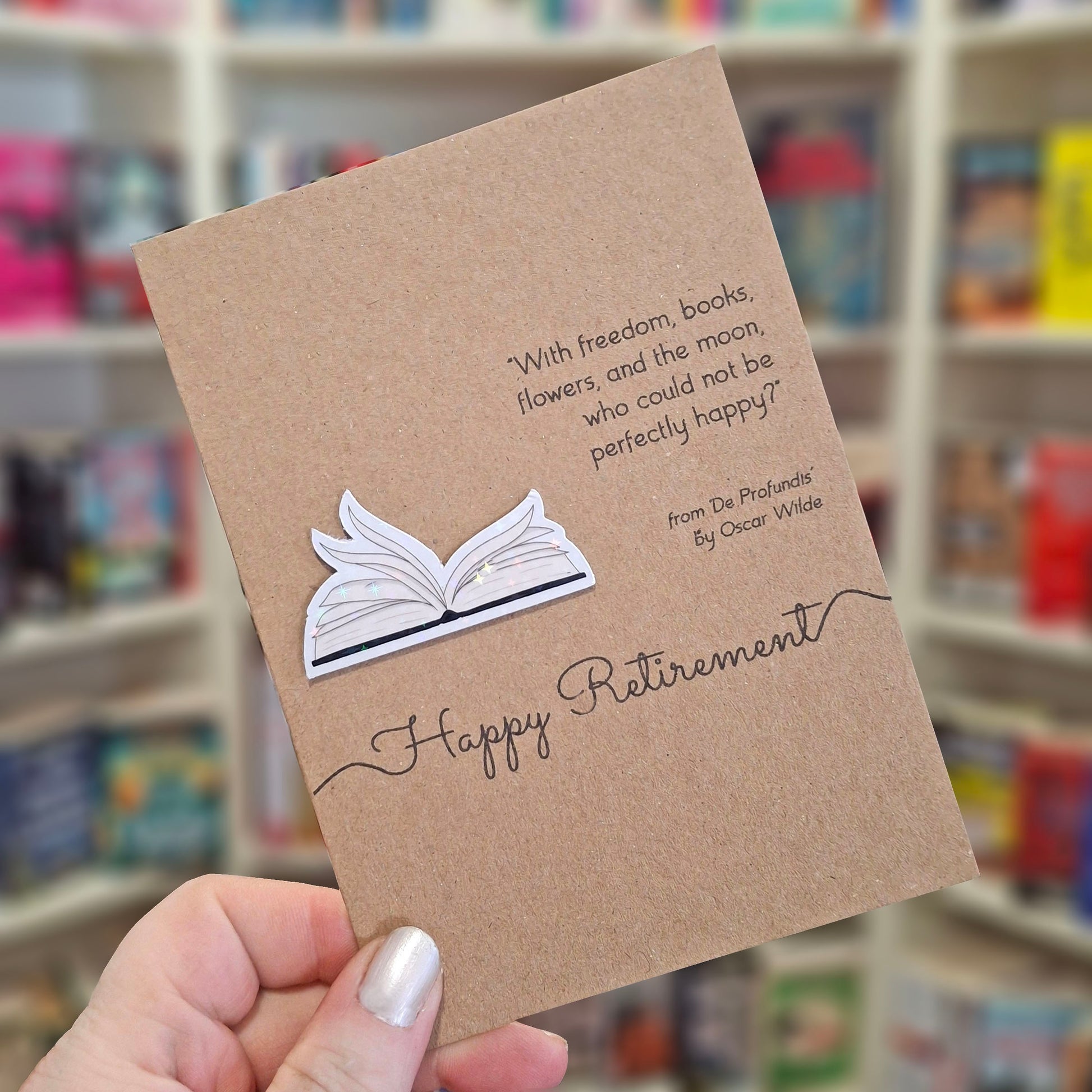 A hand holds a brown kraft happy retirement card featuring a raised holographic embellishment of an open book with gently fanned pages, and a printed quote by Oscar Wilde. The background shows a blurred bookshop interior.