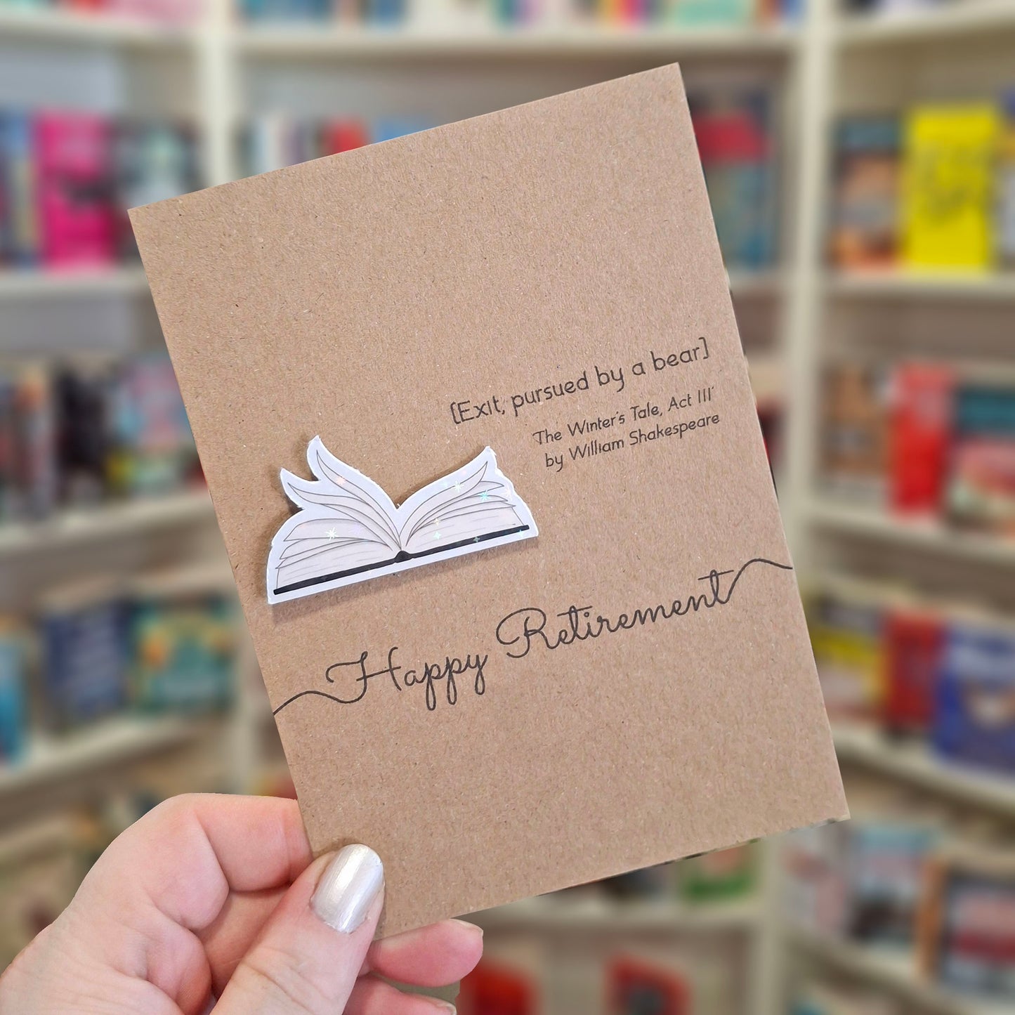 A hand holds a brown kraft retirement card featuring a raised holographic embellishment of an open book with gently fanned pages, and a printed quote by William Shakespeare. The background shows a blurred bookshop interior.