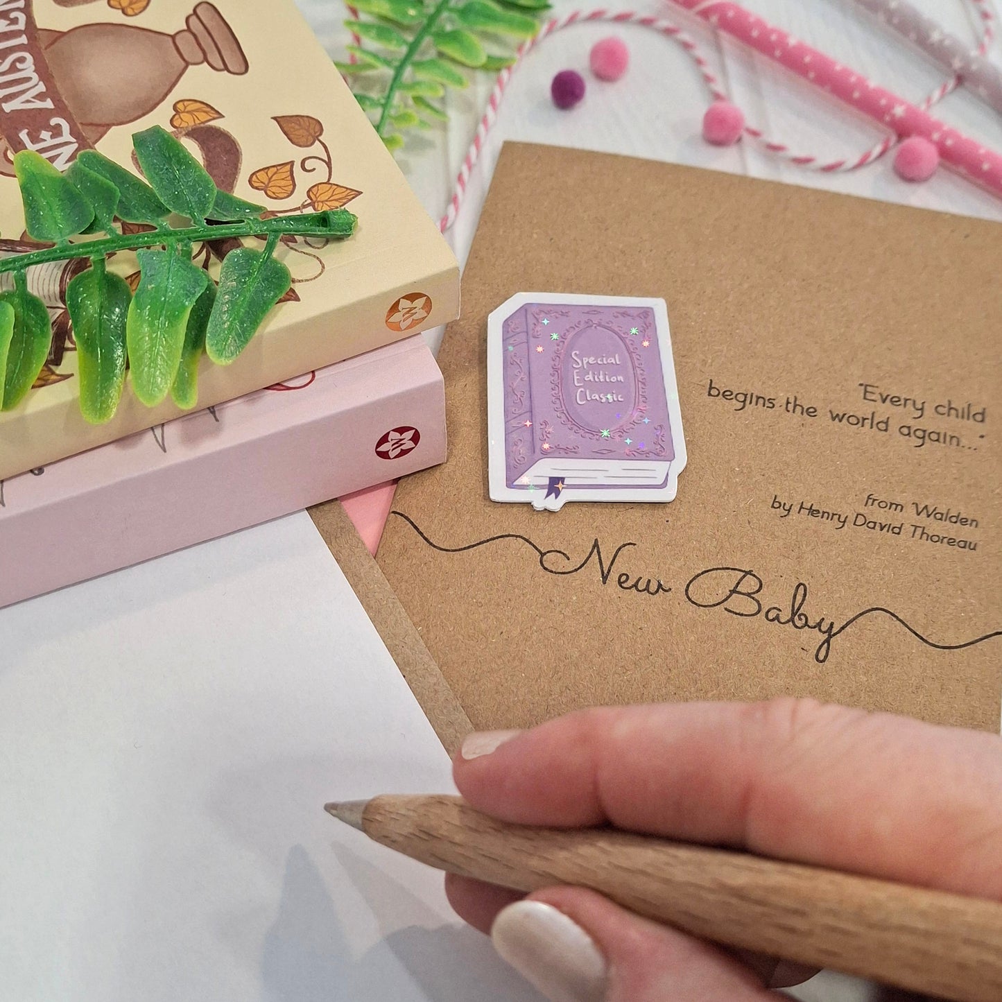 A close-up of a hand holding a wooden pen and writing a congratulations message inside a card. The card features a raised holographic embellishment of a purple special edition classic book, the words New Baby, and a quote from Walden by Henry David Thoreau.
