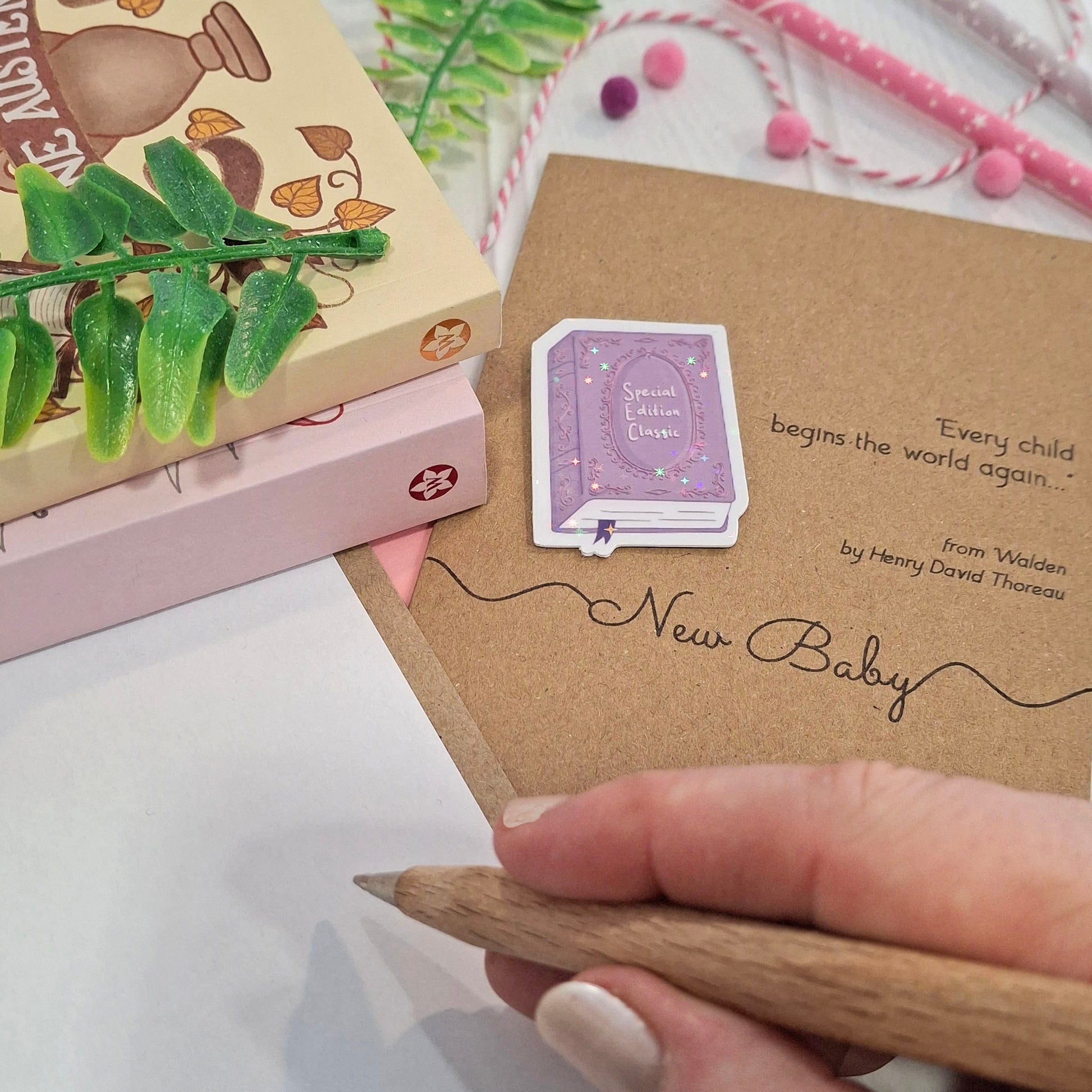 A close-up of a hand holding a wooden pen and writing a congratulations message inside a card. The card features a raised holographic embellishment of a purple special edition classic book, the words New Baby, and a quote from Walden by Henry David Thoreau.