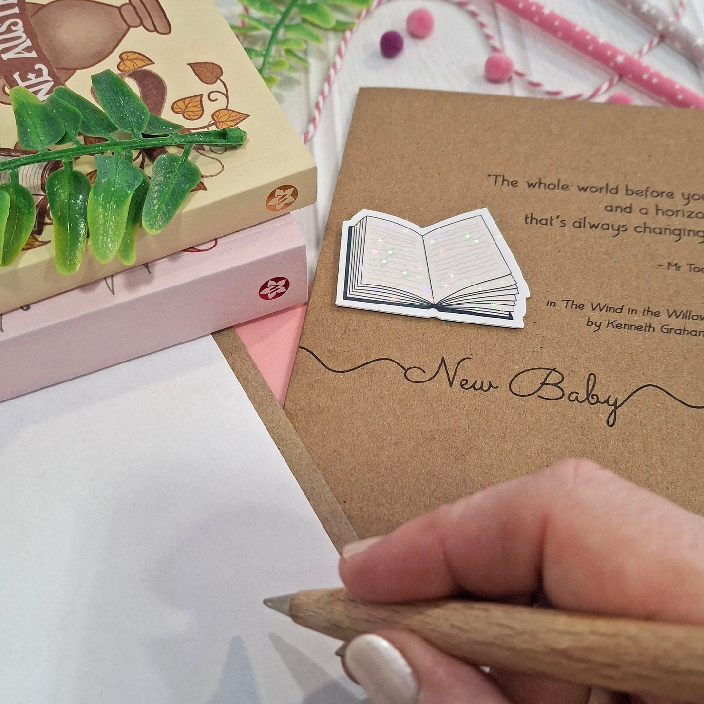 A close-up of a hand holding a wooden pen and writing inside a card. The card features a raised holographic embellishment of an open book, and reads 'New Baby', with a quote from the book The Wind in the Willows by Kenneth Grahame. 