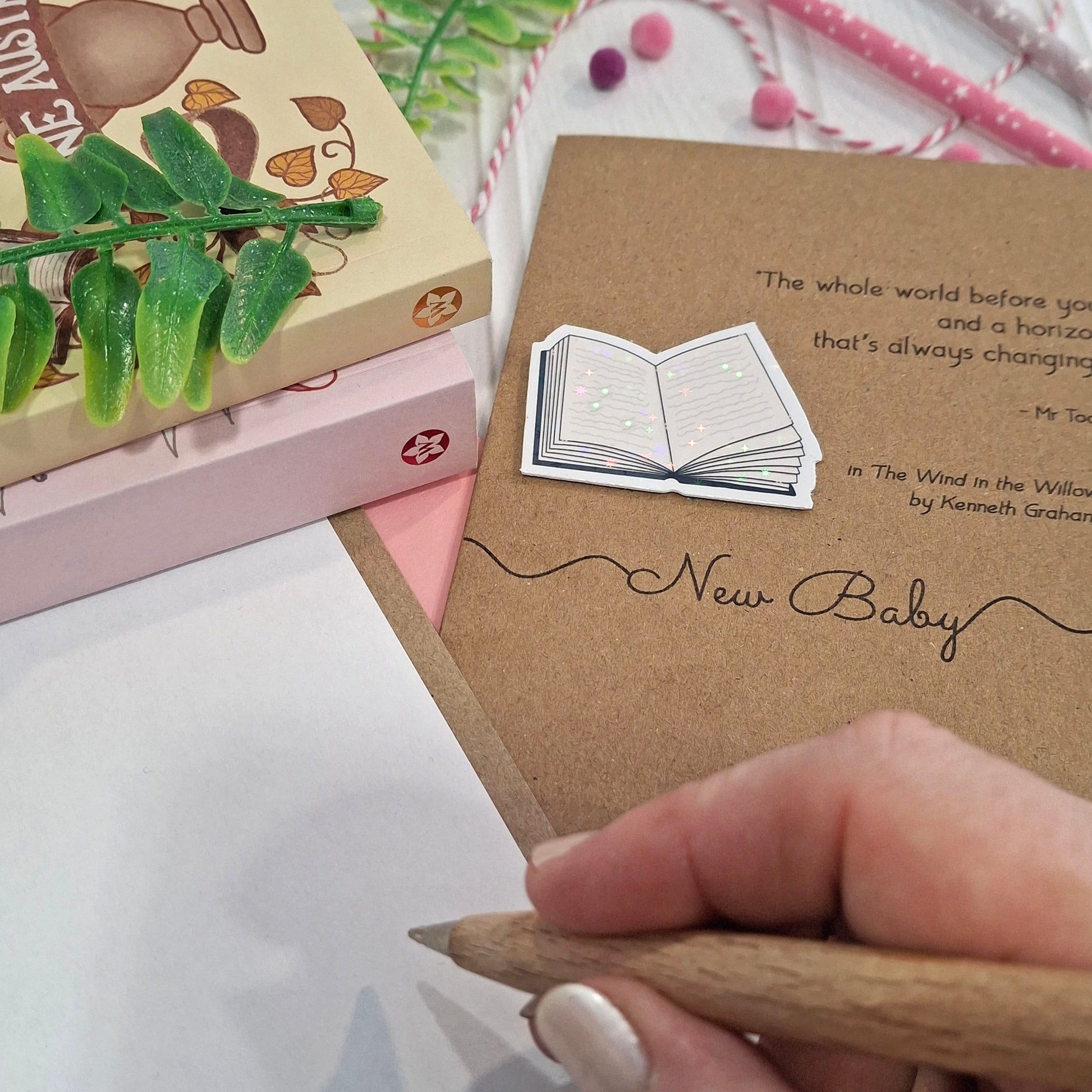 A close-up of a hand holding a wooden pen and writing inside a card. The card features a raised holographic embellishment of an open book, and reads 'New Baby', with a quote from the book The Wind in the Willows by Kenneth Grahame. 