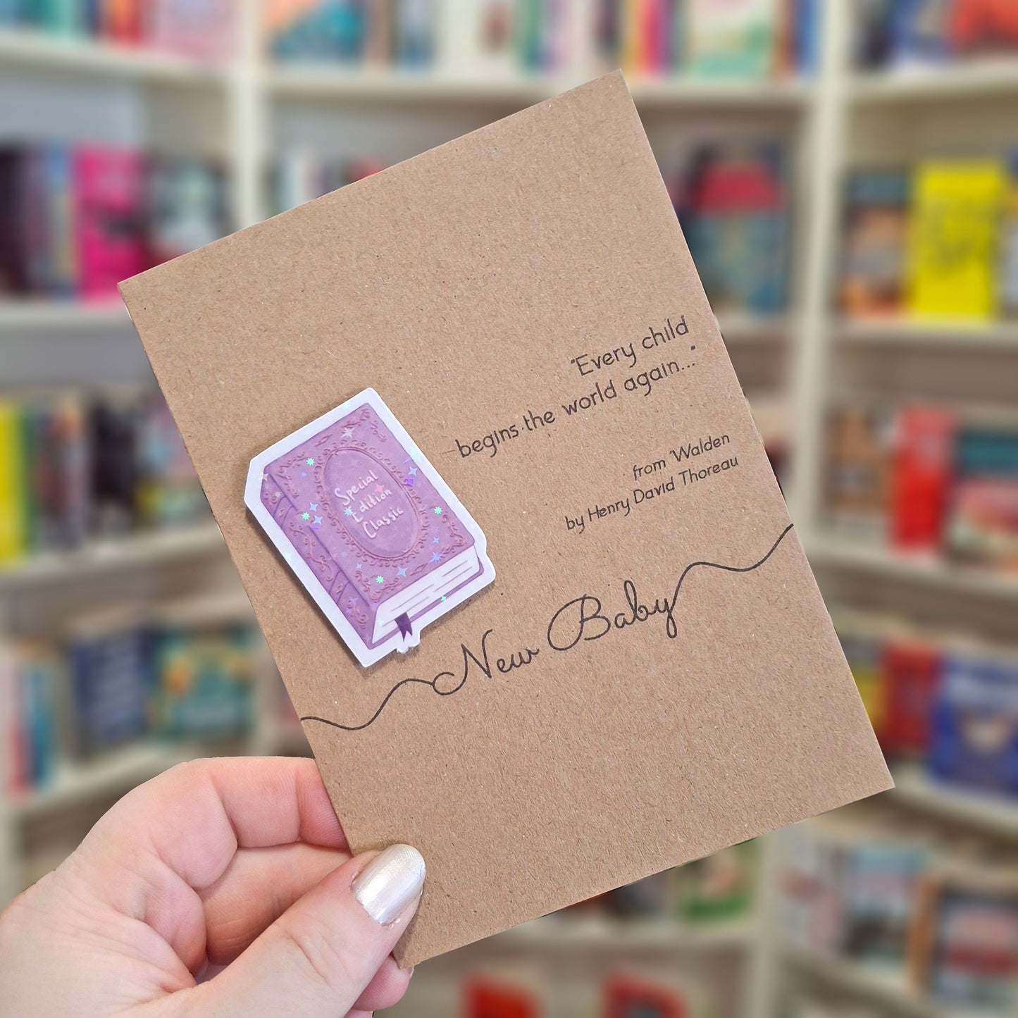 A hand holds a brown kraft new baby card featuring a raised holographic embellishment of a purple special edition classic book, and a printed quote by Henry David Thoreau. The background shows a blurred bookshop interior.