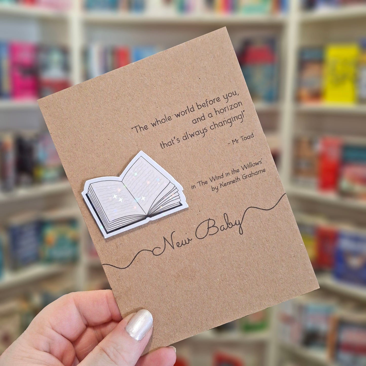 A hand holds a brown kraft new baby card featuring a raised holographic embellishment of an open book, and a printed quote by Kenneth Grahame. The background shows a blurred bookshop interior.