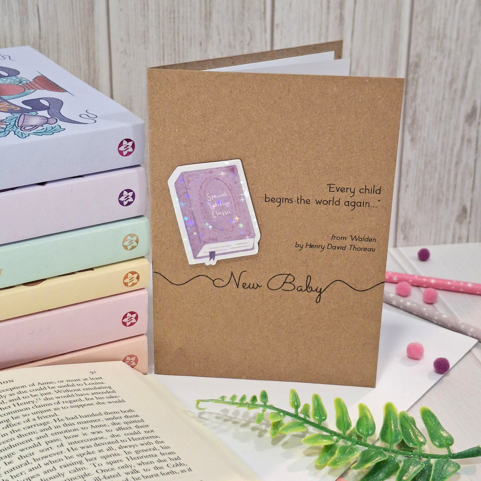 A brown kraft new baby card styled with a pastel coloured book stack, an open book and a green fern. The card features a raised holographic embellishment of a purple special edition classic book, the words New Baby, and a quote from Walden by Henry David Thoreau.