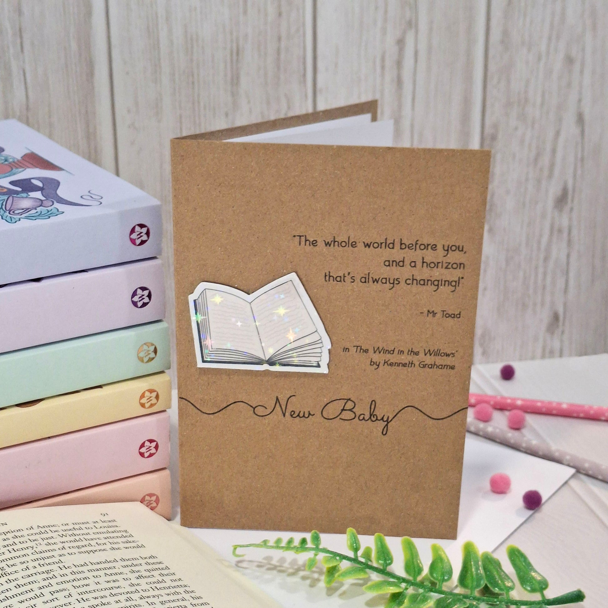 A brown kraft 'New Baby' greeting card with a raised holographic embellishment of an open book. The card reads 'New Baby', and features a quote from the book The Wind in the Willows by Kenneth Grahame. The card is styled on a tabletop with pastel book stack and foliage.