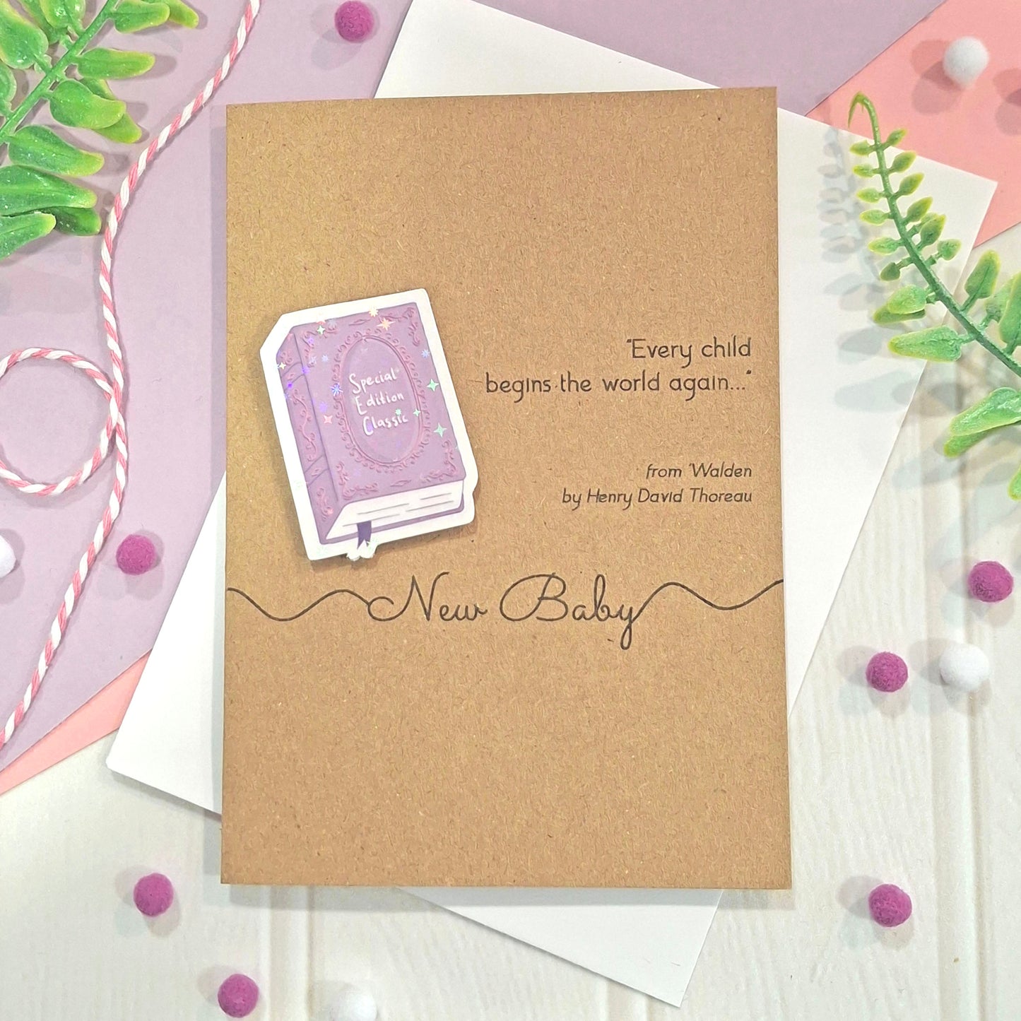 A new baby card made from brown kraft card, with a raised, holographic, illustrated story book embellishment in purple, a quote from Walden by Henry David Thoreau, and the words New Baby, all styled on a white envelope with decorative greenery and pom-poms.