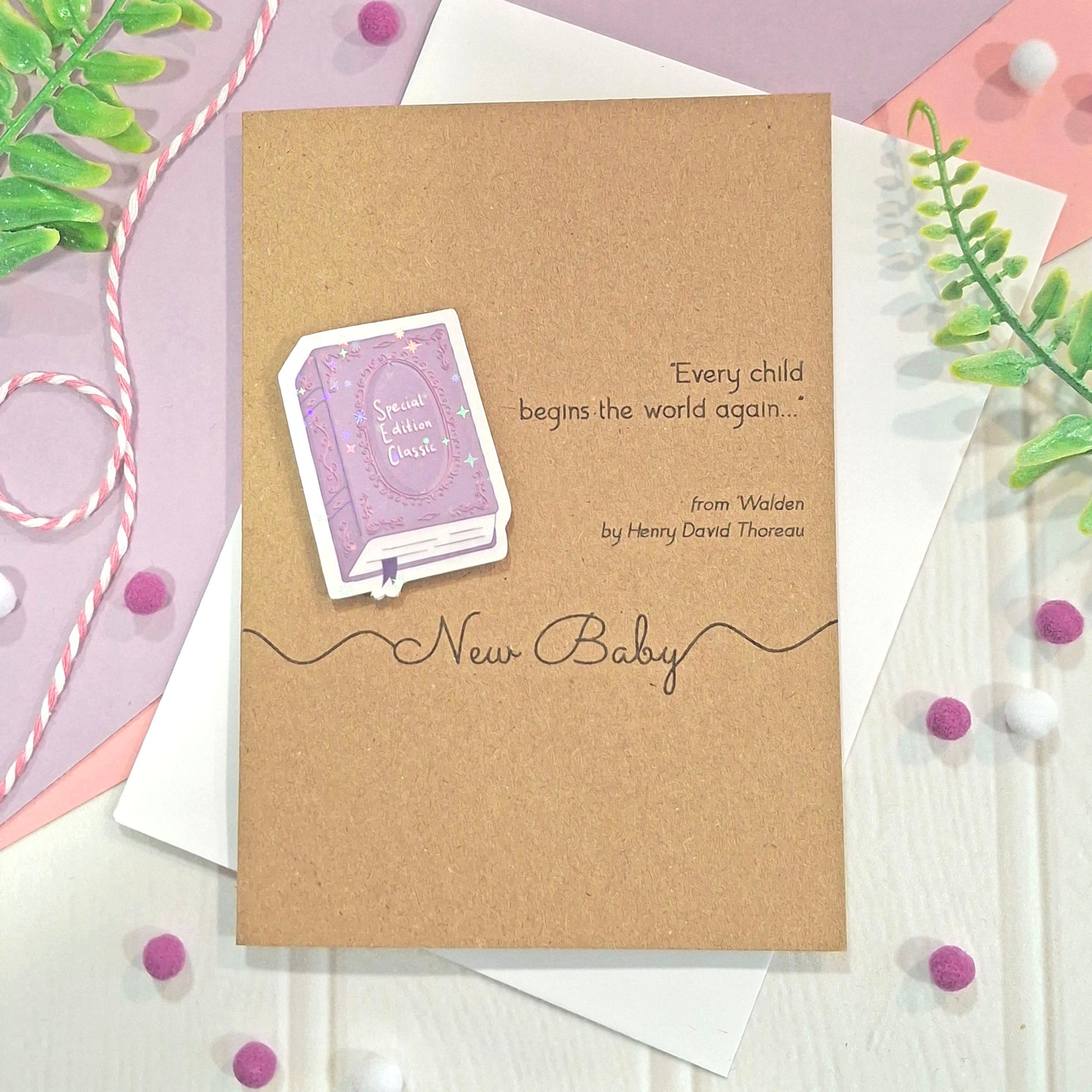 A new baby card made from brown kraft card, with a raised, holographic, illustrated story book embellishment in purple, a quote from Walden by Henry David Thoreau, and the words New Baby, all styled on a white envelope with decorative greenery and pom-poms.