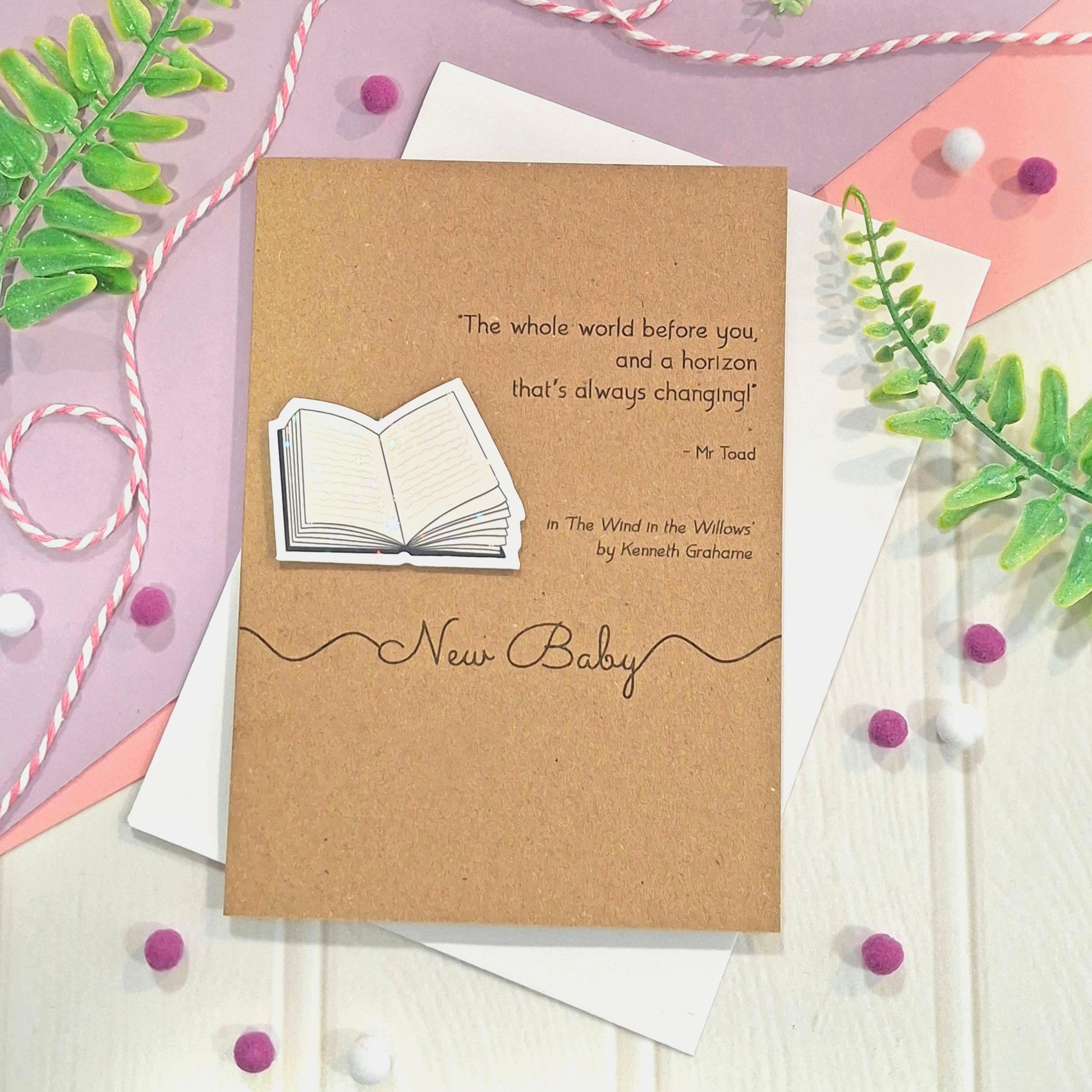 A kraft new baby greeting card with a quote from the book 'The Wind in the Willows' by Kenneth Grahame, a raised, holographic, illustrated open book embellishment, and the words 'New Baby' in elegant script, all styled with white envelopes, green leaves, pink and white ribbons, and small pom-poms.