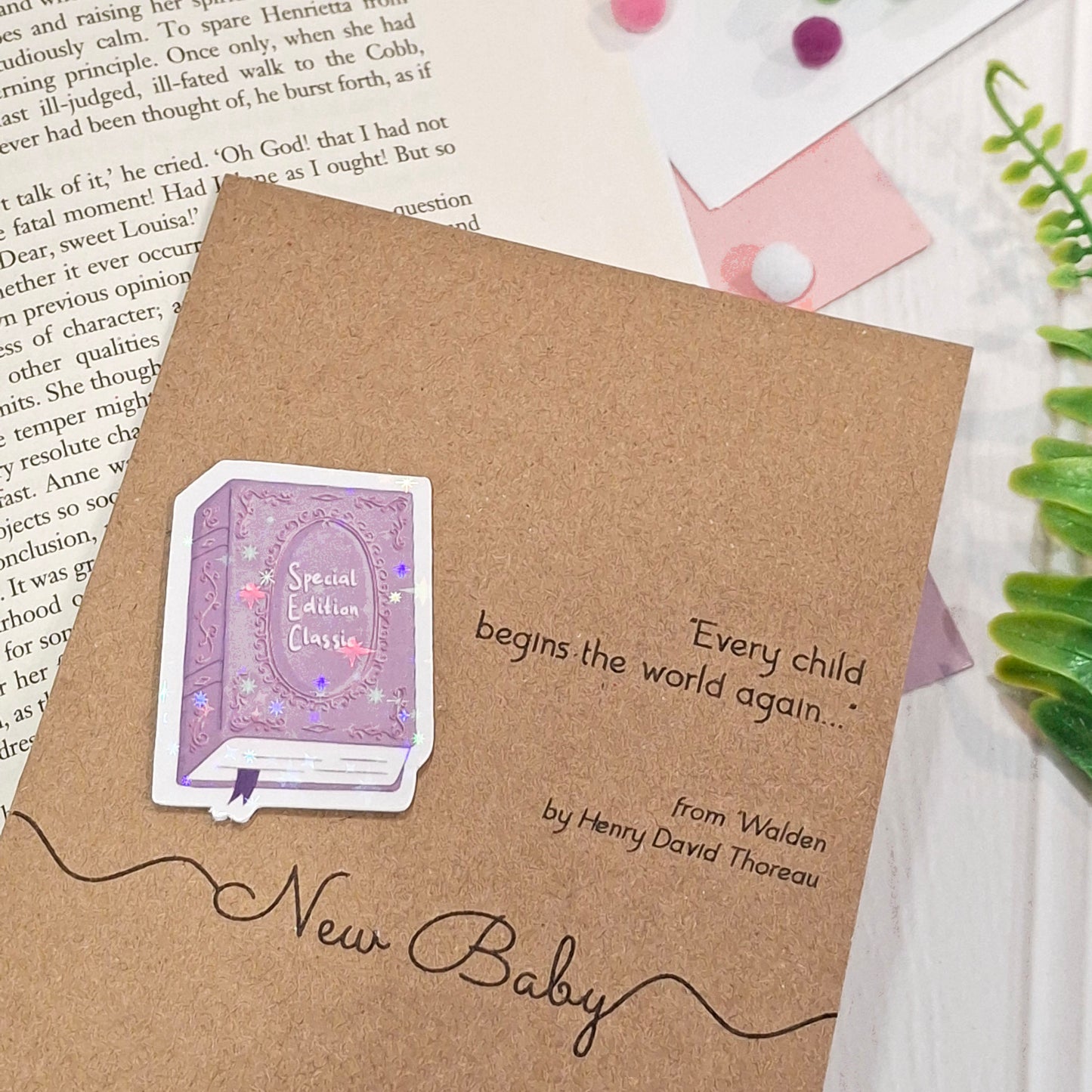 A kraft New Baby card styled on top of an open book with pink and green decorative accents. The card features a raised holographic embellishment of a purple special edition classic book, the words New Baby, and a quote from Walden by Henry David Thoreau.