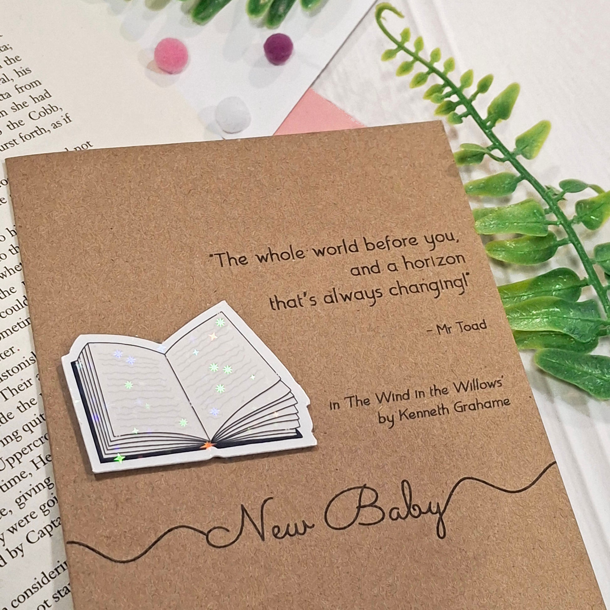 A brown kraft 'New Baby' greeting card with a raised holographic embellishment of an open book. The card reads 'New Baby', and features a quote from the book The Wind in the Willows by Kenneth Grahame. The card is styled on an open book page surrounded with greenery.