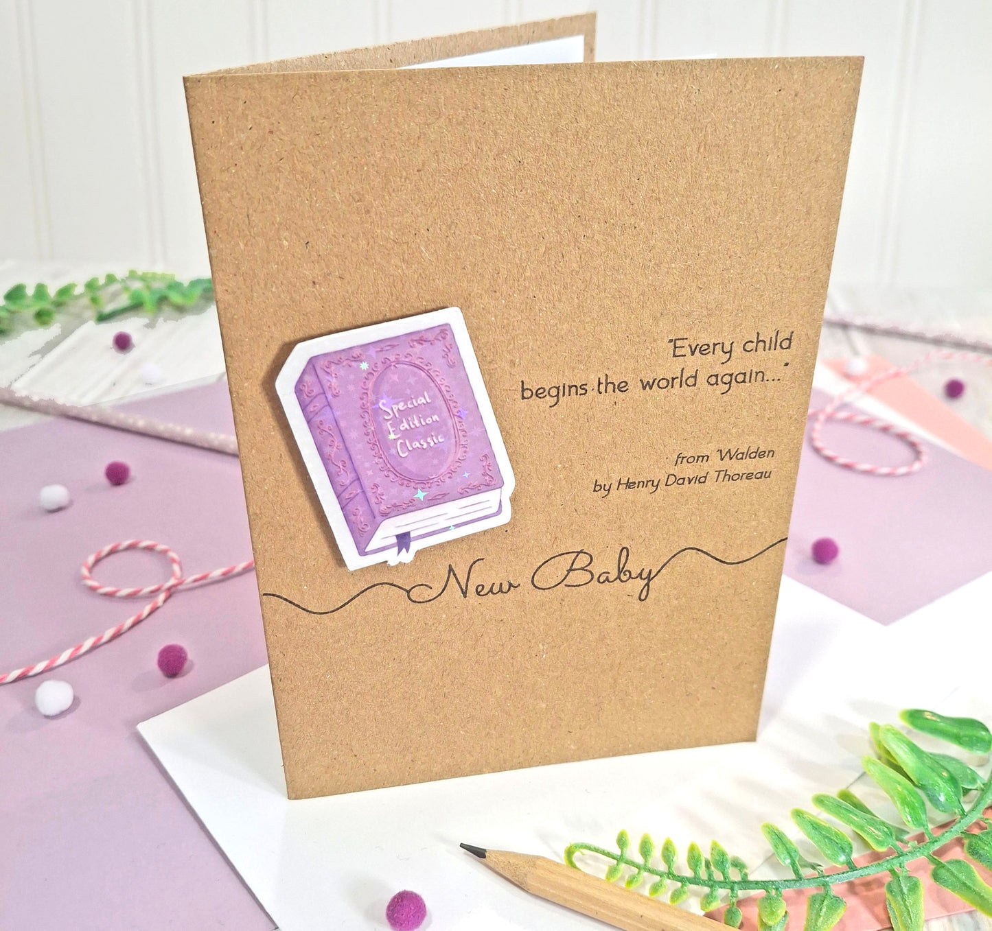 A kraft card for a new baby, featuring a raised, holographic, illustrated story book embellishment in purple, a quote from Walden by Henry David Thoreau, and the words 'New Baby', all styled on a white and pastel background.