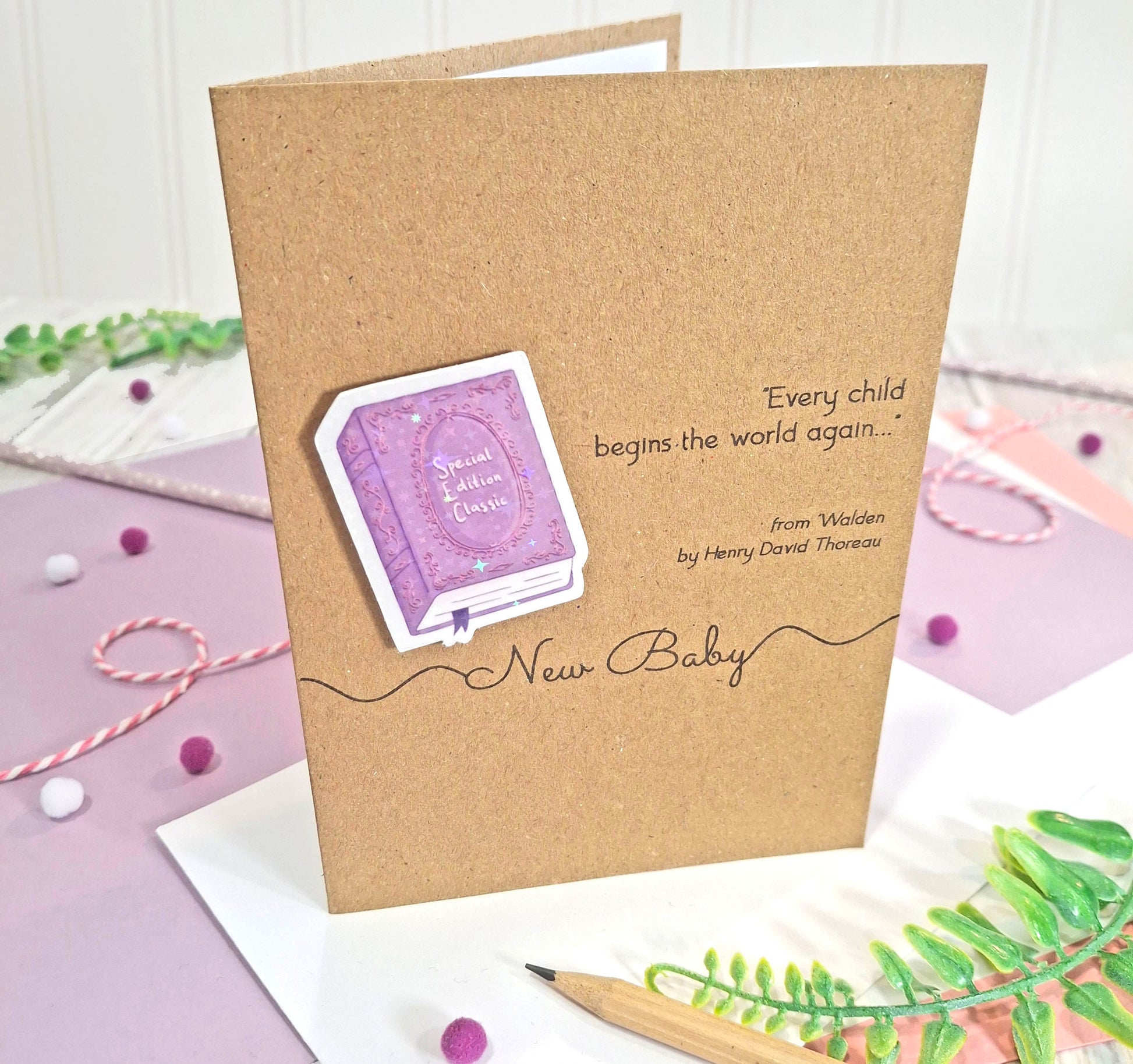 A kraft card for a new baby, featuring a raised, holographic, illustrated story book embellishment in purple, a quote from Walden by Henry David Thoreau, and the words 'New Baby', all styled on a white and pastel background.