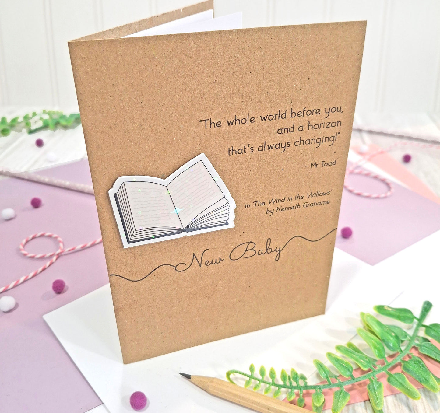 A kraft card for a 'New Baby' featuring a printed quote from the book The Wind in the Willows by Kenneth Grahame, and a raised, holographic, illustrated open book embellishment, all displayed on a styled pastel background with a pencil and decorative elements.