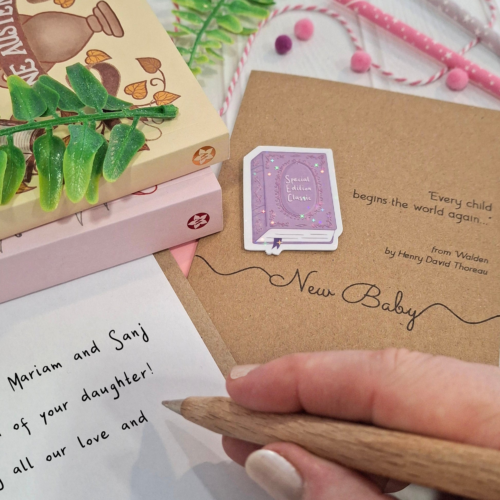 A close-up of a hand holding a wooden pen and writing inside a card. The card features a raised holographic embellishment of a purple special edition classic book, the words New Baby, and a quote from Walden by Henry David Thoreau.