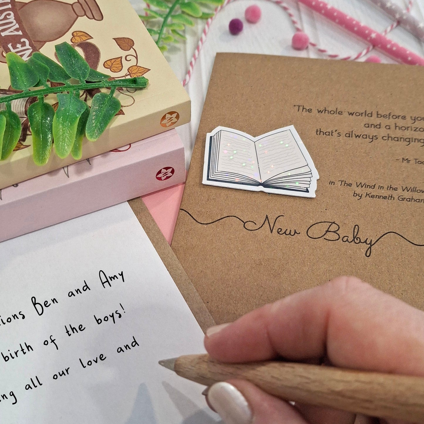 A close-up of a hand holding a wooden pen and writing a congratulations on your new baby message inside a card. The card features a raised holographic embellishment of an open book, and reads 'New Baby', with a quote from the book The Wind in the Willows by Kenneth Grahame. 