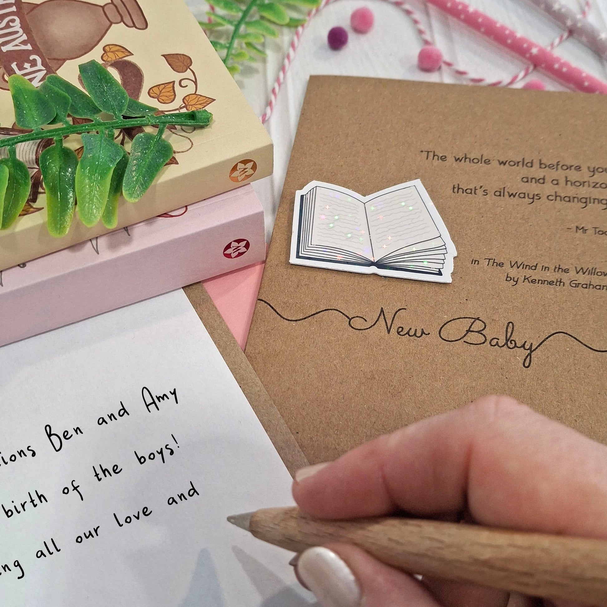 A close-up of a hand holding a wooden pen and writing a congratulations on your new baby message inside a card. The card features a raised holographic embellishment of an open book, and reads 'New Baby', with a quote from the book The Wind in the Willows by Kenneth Grahame. 