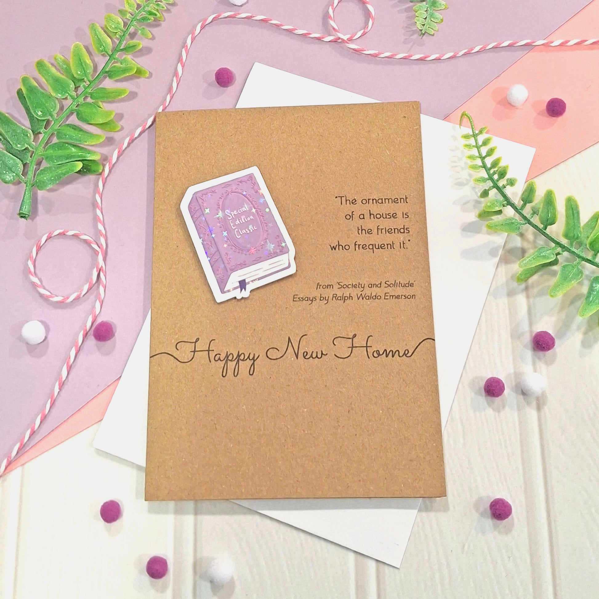 A handmade 'Happy New Home' greeting card made from brown kraft card, decorated with a raised, holographic, illustrated story book embellishment in purple, a quote from Society and Solitude by Ralph Waldo Emerson, and the words 'Happy New Home'. The card is styled in a flat-lay arrangement with white envelopes, pastel backgrounds, green leaves, and pink-and-white string.