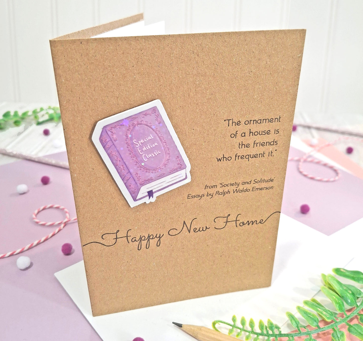 A kraft brown greeting card with  a raised, holographic, illustrated story book embellishment in purple, a quote from Society and Solitude by Ralph Waldo Emerson, and the words 'Happy New Home' in script. The card is displayed in a styled flat-lay setting with decorative props.