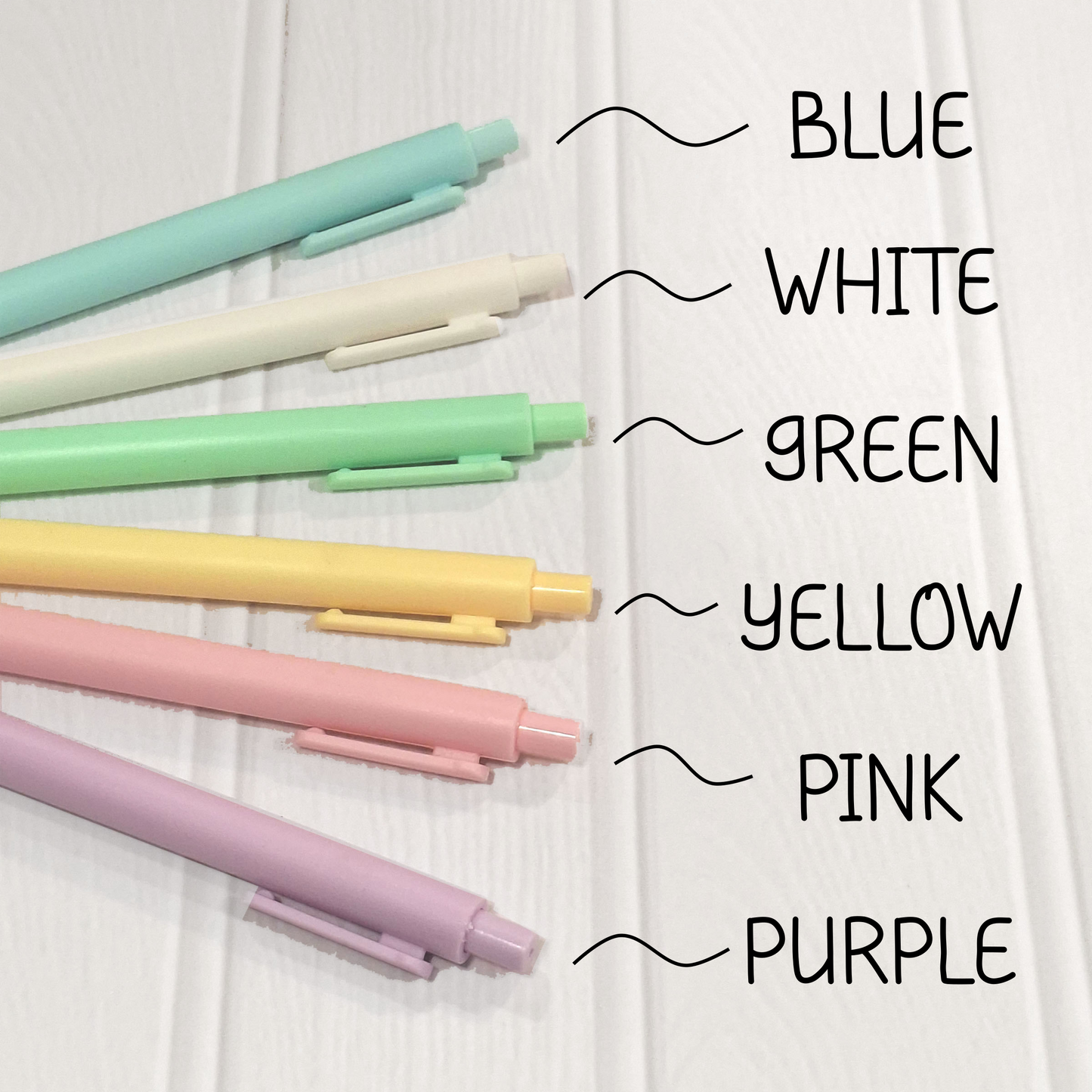 Six pastel-coloured pens arranged on a white wood background, with handwritten labels pointing to each colour: BLUE, WHITE, GREEN, YELLOW, PINK, and PURPLE.