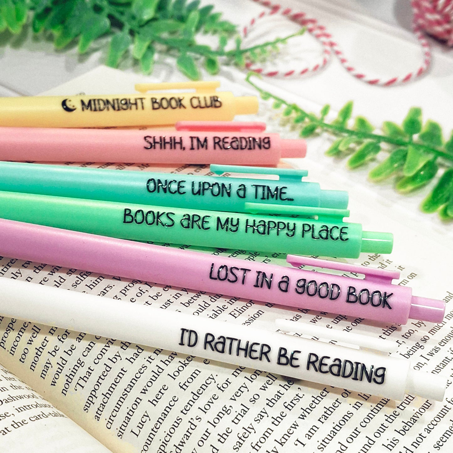 Six colourful pens with book-themed quotes are arranged diagonally on an open book with decorative green leaves and a pink-and-white string in the background. The pens are yellow, pink, blue, green, purple, and white.