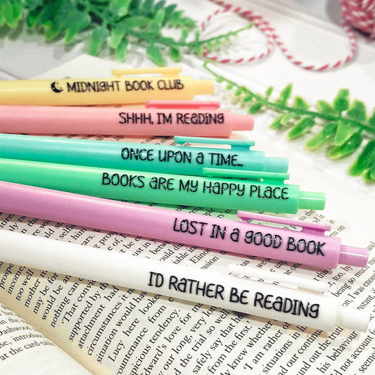 Six colourful pens with book-themed quotes are arranged diagonally on an open book with decorative green leaves and a pink-and-white string in the background. The pens are yellow, pink, blue, green, purple, and white.