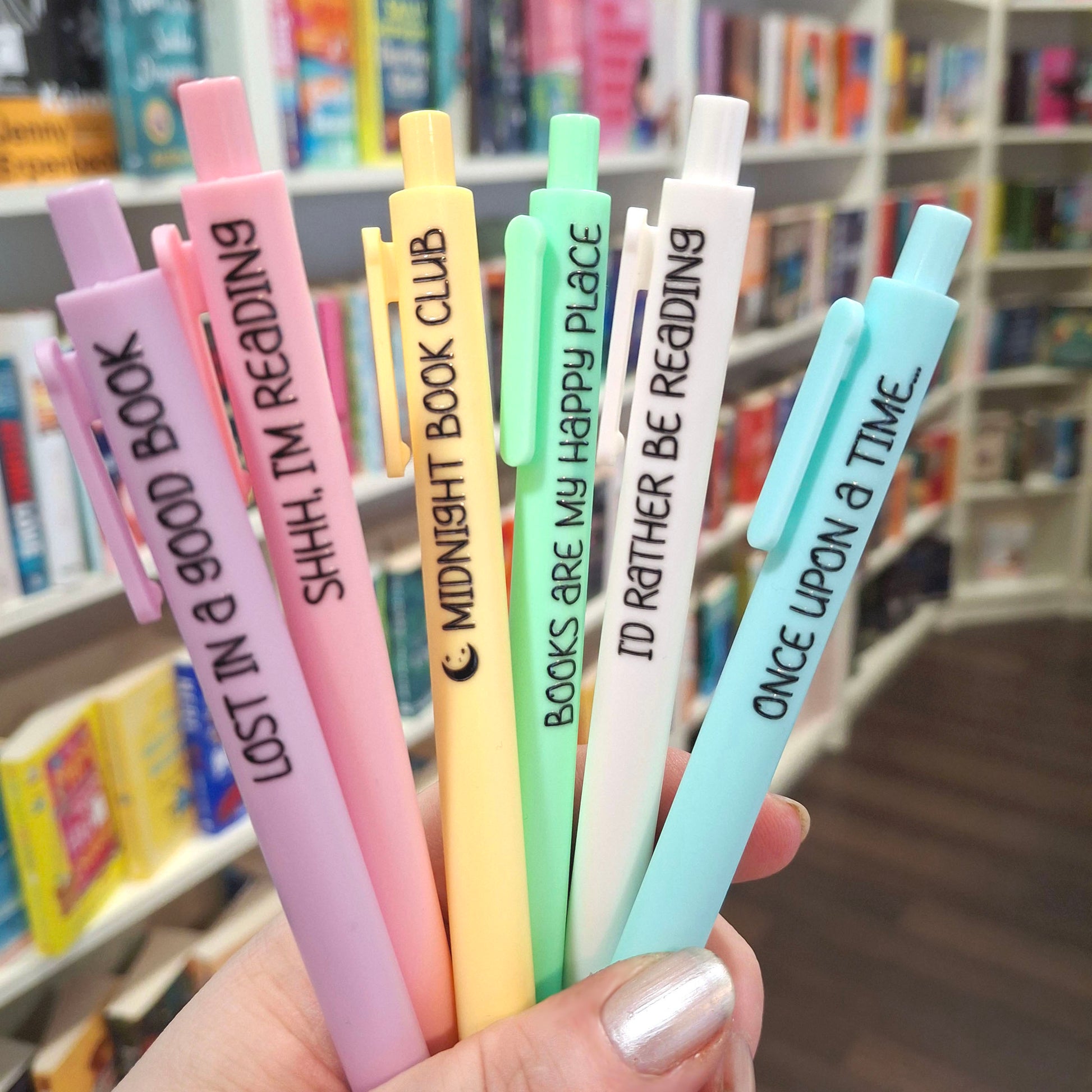 A hand holding six pastel-coloured pens with book-themed quotes printed in black text, set against a blurred background of a bookshelf.