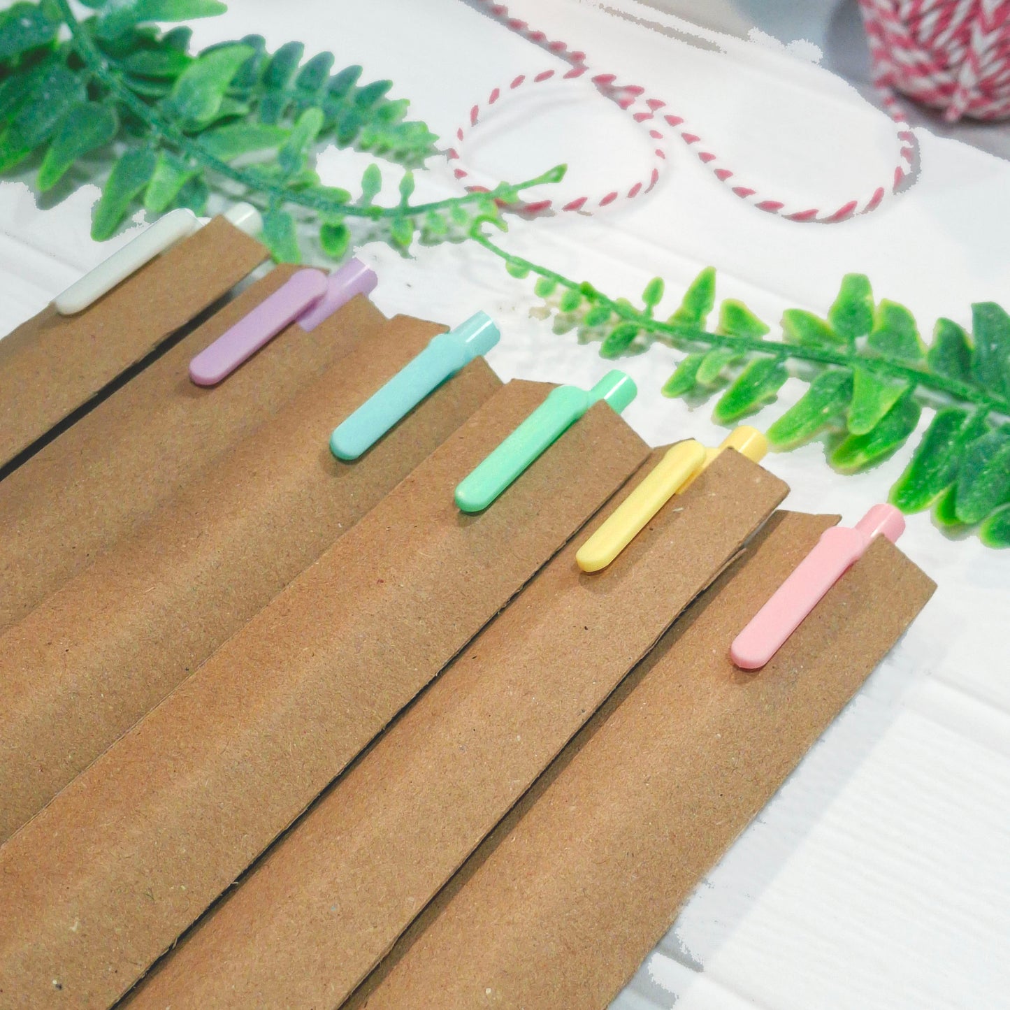 Six pastel-coloured pens in pink, yellow, blue, green, and purple, packaged in kraft sleeves, arranged on a white surface with decorative green leaves and a spool of pink-and-white twine.