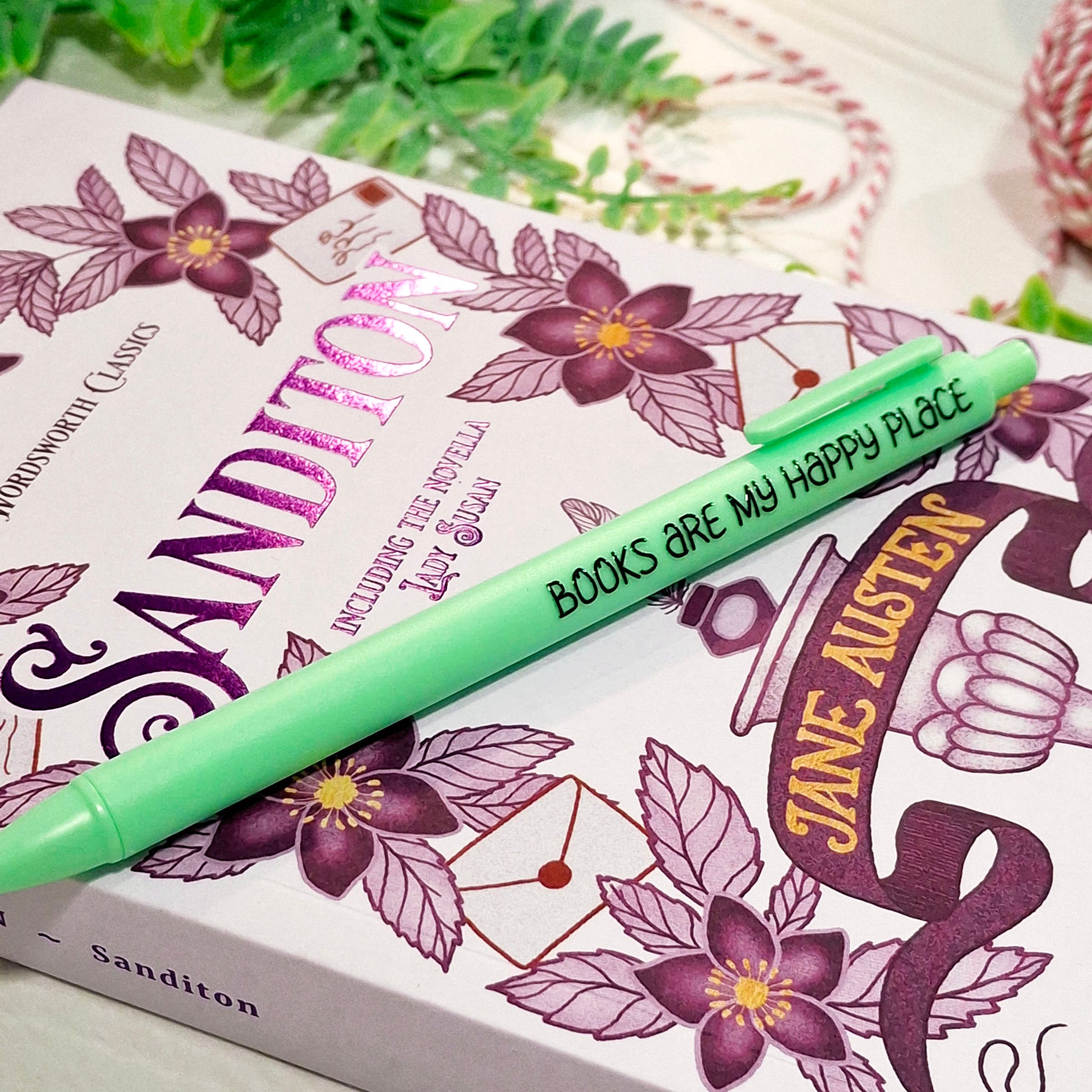 A green pen with the printed phrase 'BOOKS ARE MY HAPPY PLACE' resting diagonally across a book cover featuring purple floral illustrations and Jane Austen-themed artwork.