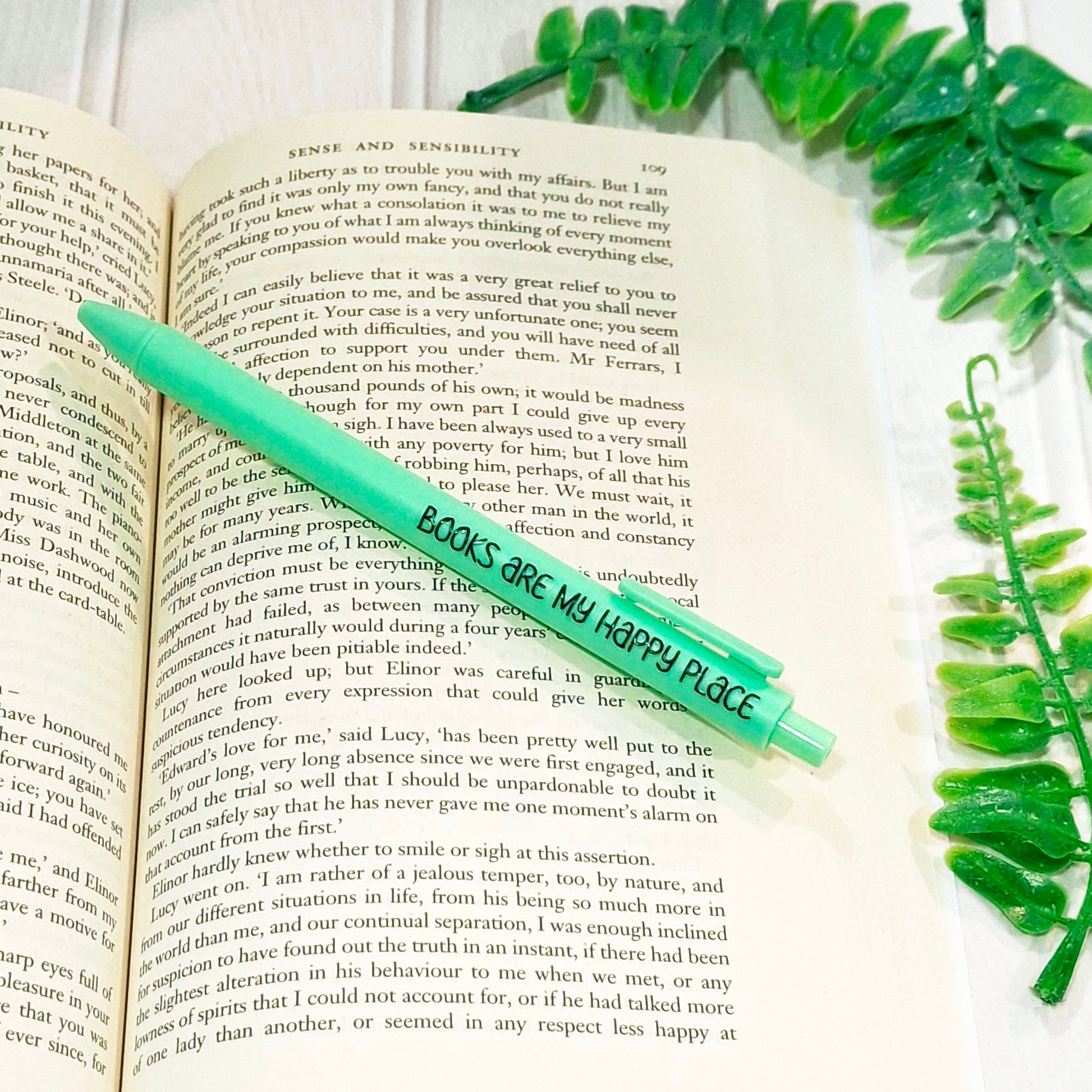 A green pen with the printed phrase 'BOOKS ARE MY HAPPY PLACE' resting diagonally across an open book.