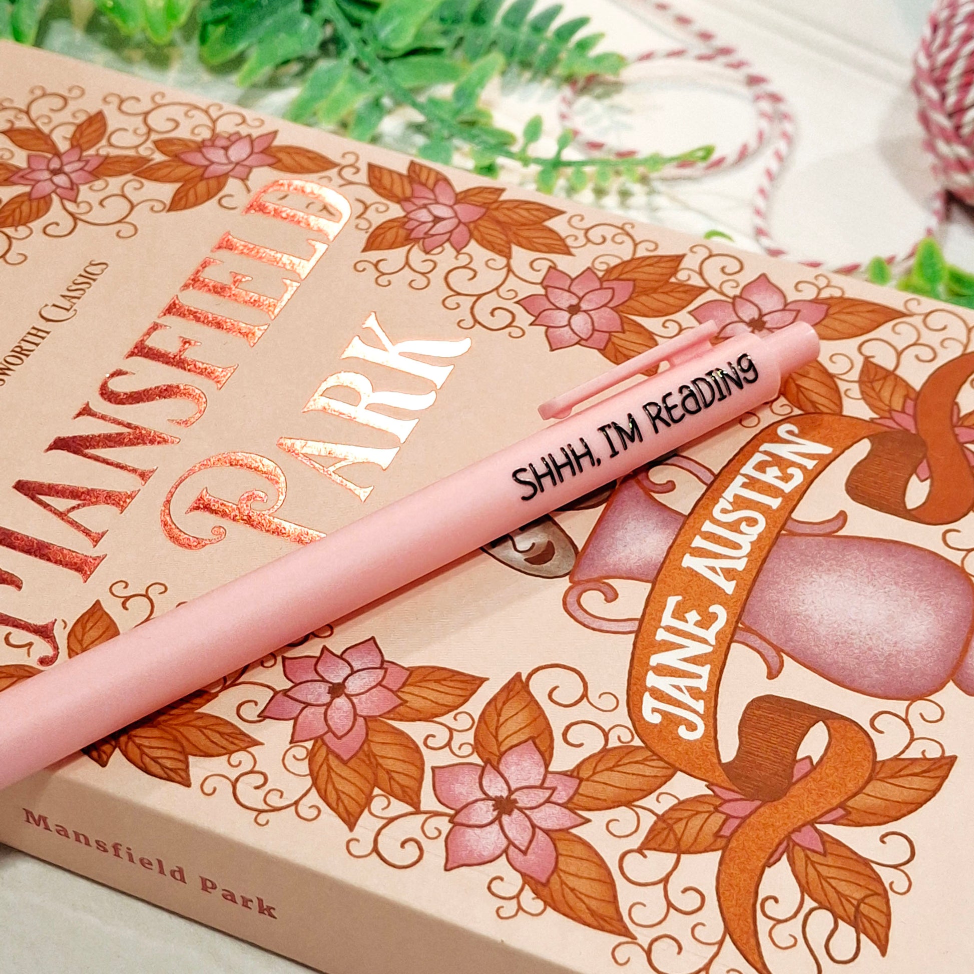 A light pink pen with the text 'SHHH, I'M READING' in black lettering, resting diagonally across a Jane Austen-themed book cover featuring gold foil lettering and floral illustrations.