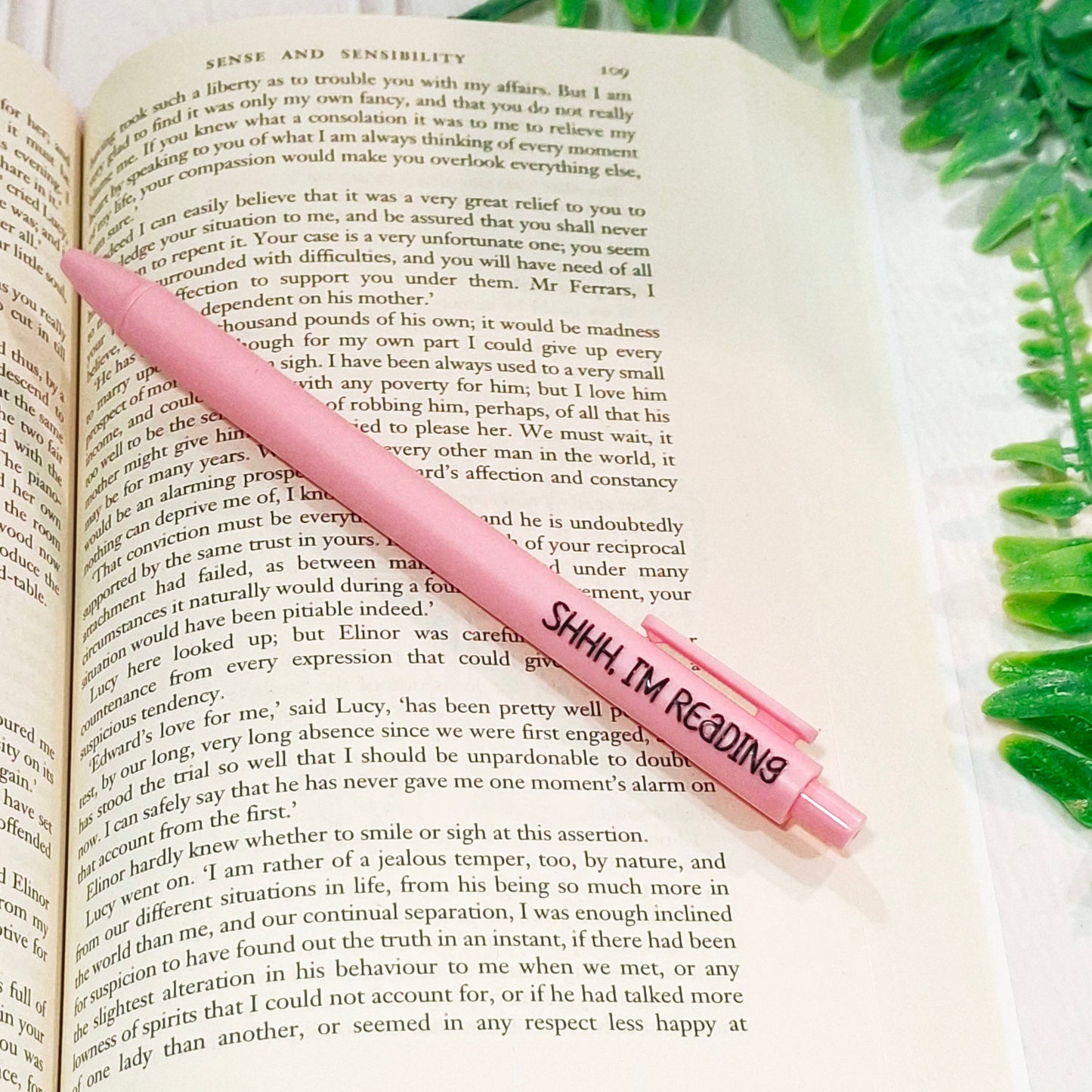 A light pink pen with the text 'SHHH, I'M READING' in black lettering, resting diagonally across an open book page.