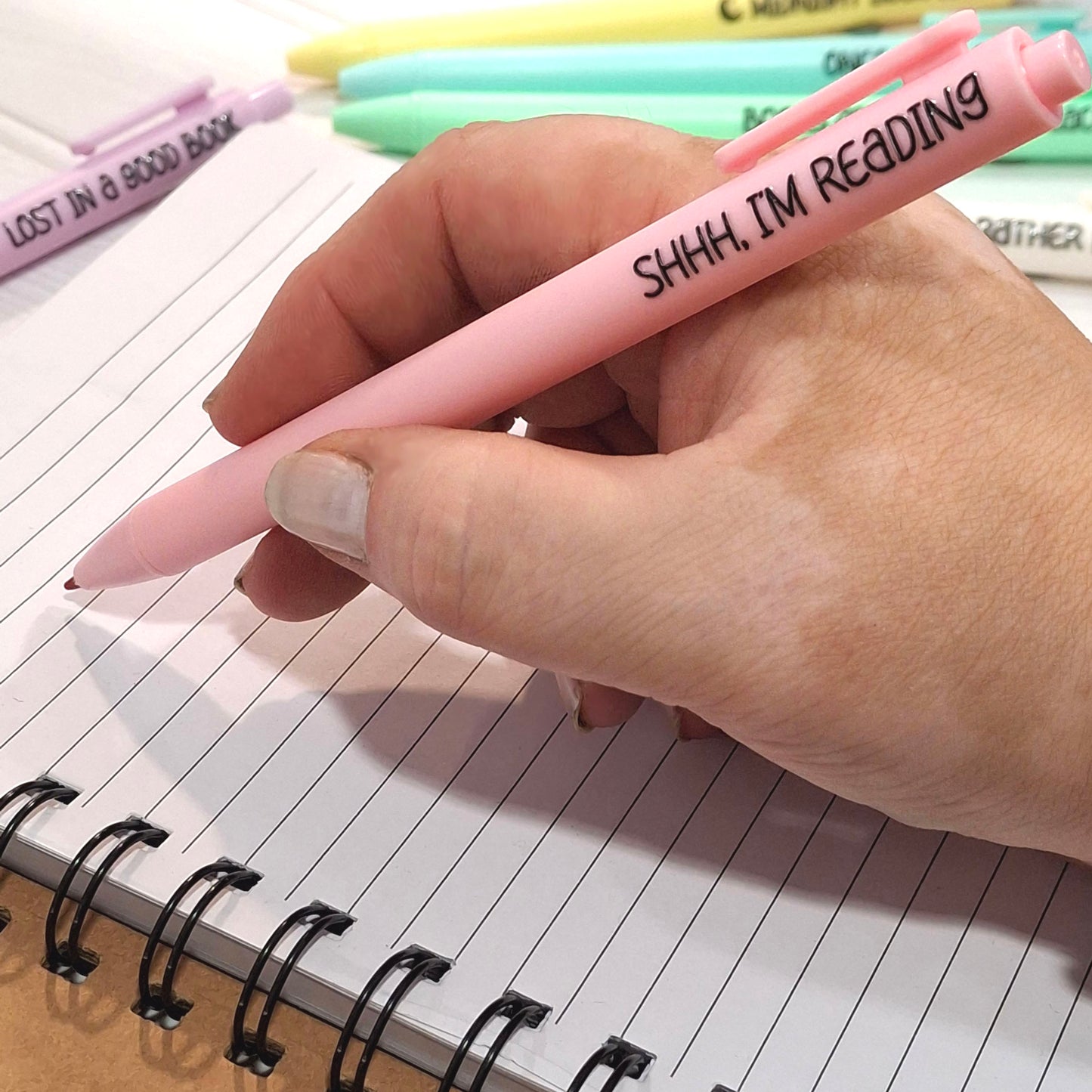 A hand holding a pink retractable pen with the text 'SHHH, I'M READING' in black lettering, poised over an open spiral notebook.