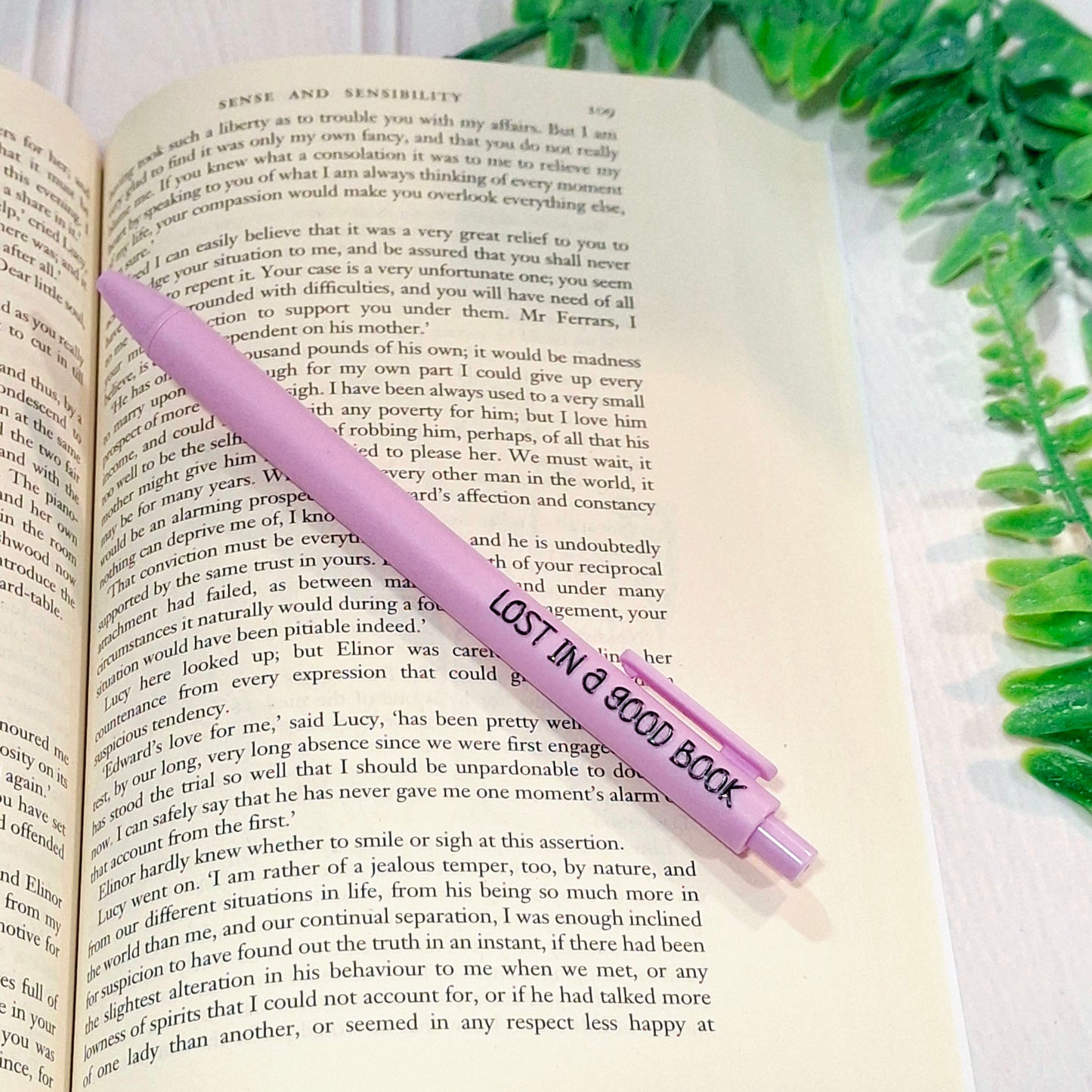 A light purple pen with black text reading 'LOST IN A GOOD BOOK' resting on a an open book page.