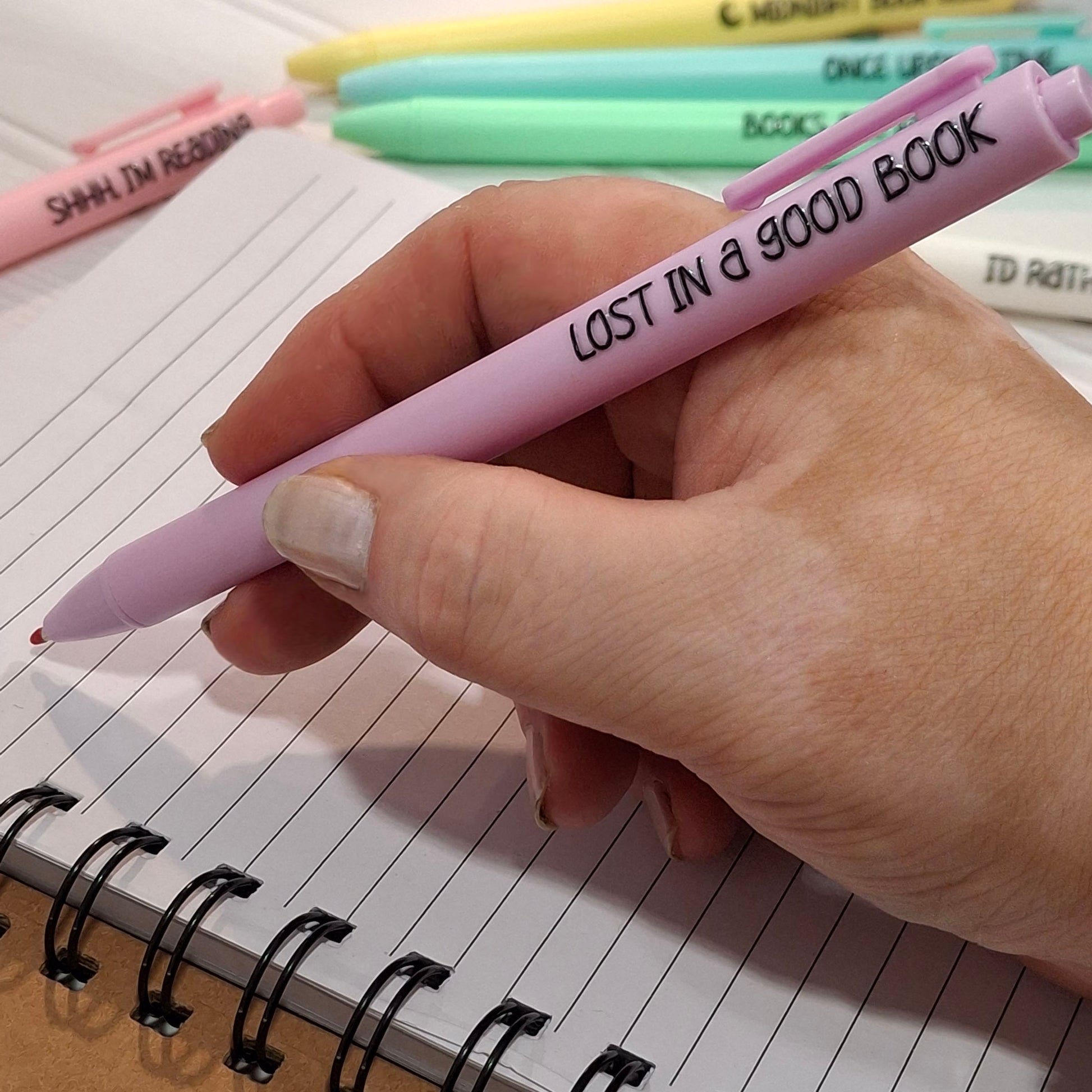 A hand holding a purple pen with the phrase 'LOST IN A GOOD BOOK' printed in black. The pen is shown above an open spiral notebook, with other colourful pens blurred in the background.
