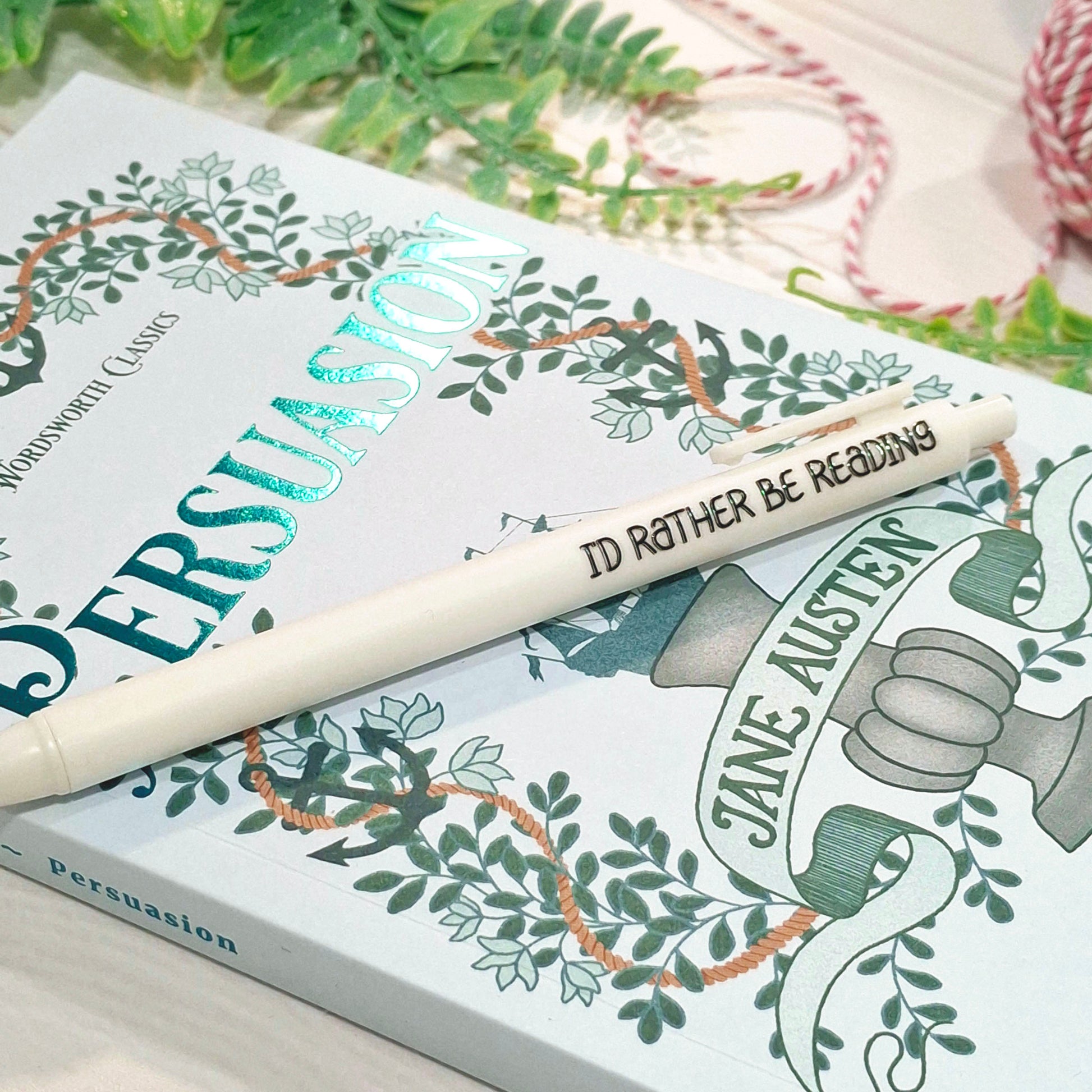 A white pen with 'I'd Rather Be Reading' printed on it, resting diagonally across a blue illustrated book titled 'Persuasion'.