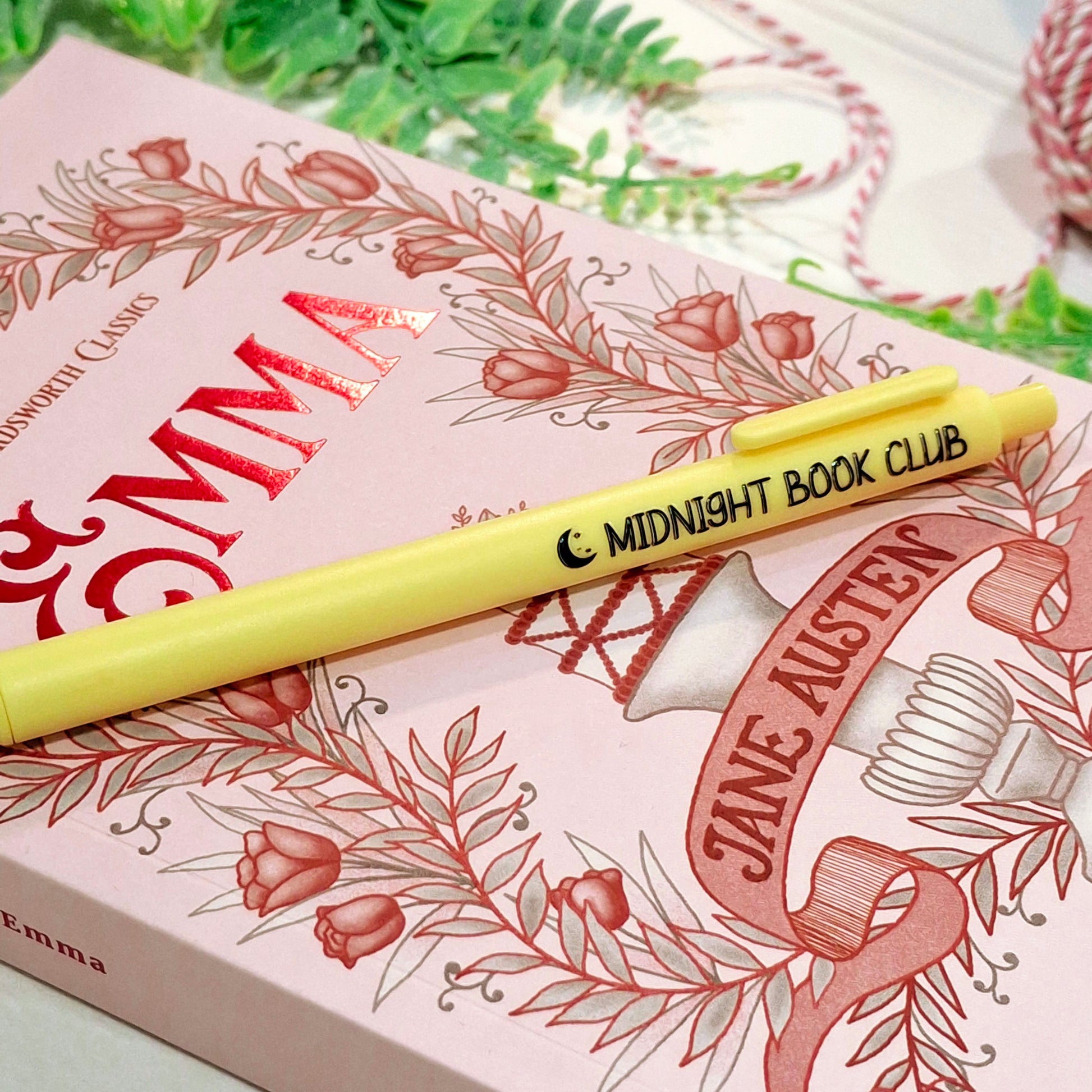 A yellow pen with 'MIDNIGHT BOOK CLUB' printed on it, resting diagonally across a pink illustrated book titled 'Emma'.